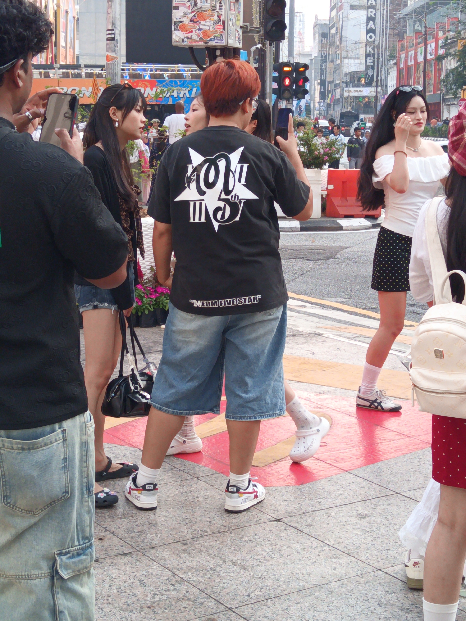 In a street scene in Kuala Lumpur, Malaysia, several young people are gathered on a tiled sidewalk. A person on the left, wearing a black patterned shirt and light blue denim cargo pants, holds a smartphone up, seemingly taking a photo. To their right, a young woman with long dark hair and sunglasses perched on her head, wears a white off-the-shoulder top with ruffles and a black skirt with white polka dots. She is looking to her right and touching her hair. Next to her, a person with bright red hair, wearing a black t-shirt with a white graphic and denim shorts, is also looking to their right. They are standing in front of a flower display. In the background, a busy street with buildings and traffic is visible, including traffic lights and a sign for "KARAOKE." Another young woman is visible on the far right, wearing a white top and a red skirt with white polka dots, carrying a white backpack. The overall atmosphere appears casual and social.