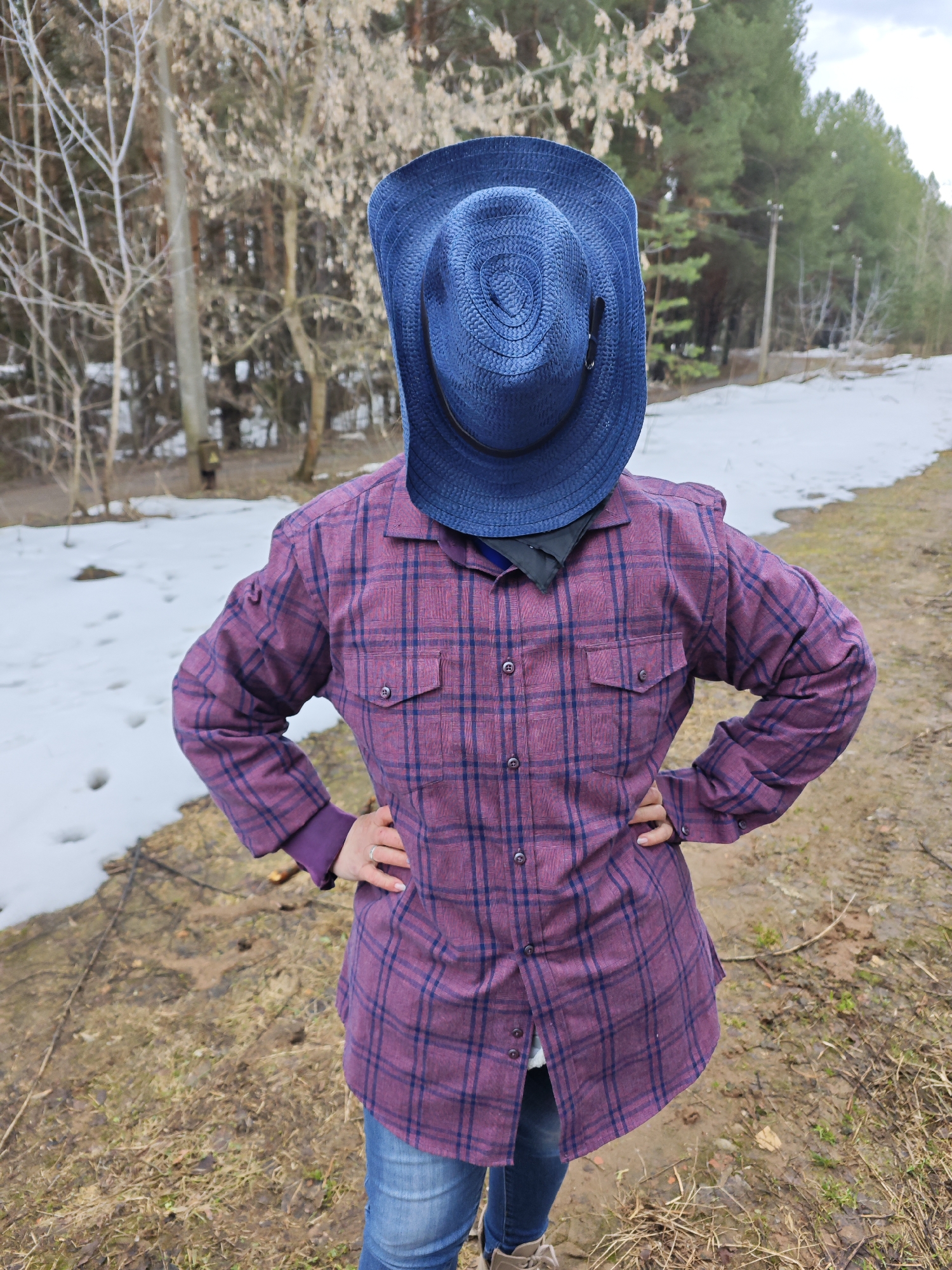 A person stands in a wooded area near Chaikovsky, Russia, wearing a purple and navy plaid shirt, jeans, and boots. A blue straw hat covers their face. The person's hands are on their hips. Patches of snow are visible on the ground to the left, with trees and a dirt path in the background.