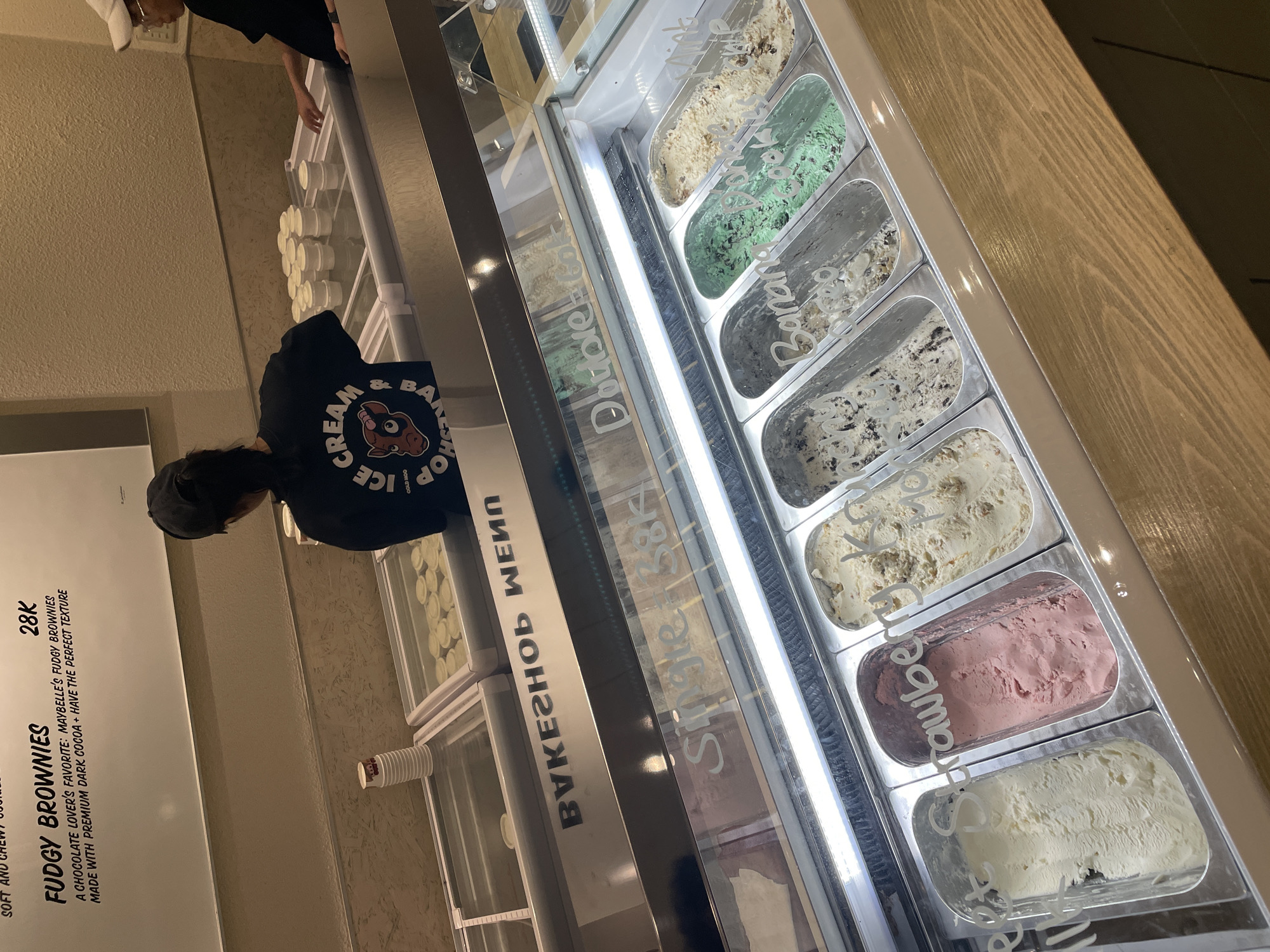 A person in a black shirt and hat stands in front of a display case of ice cream at a bakery in North Jakarta, Indonesia. The ice cream flavors include mint chocolate chip, banana, cookies and cream, and strawberry. A sign advertises "Fudgy Brownies" for 28K. The bakery's name appears to be "Cream & Bake Shop."
