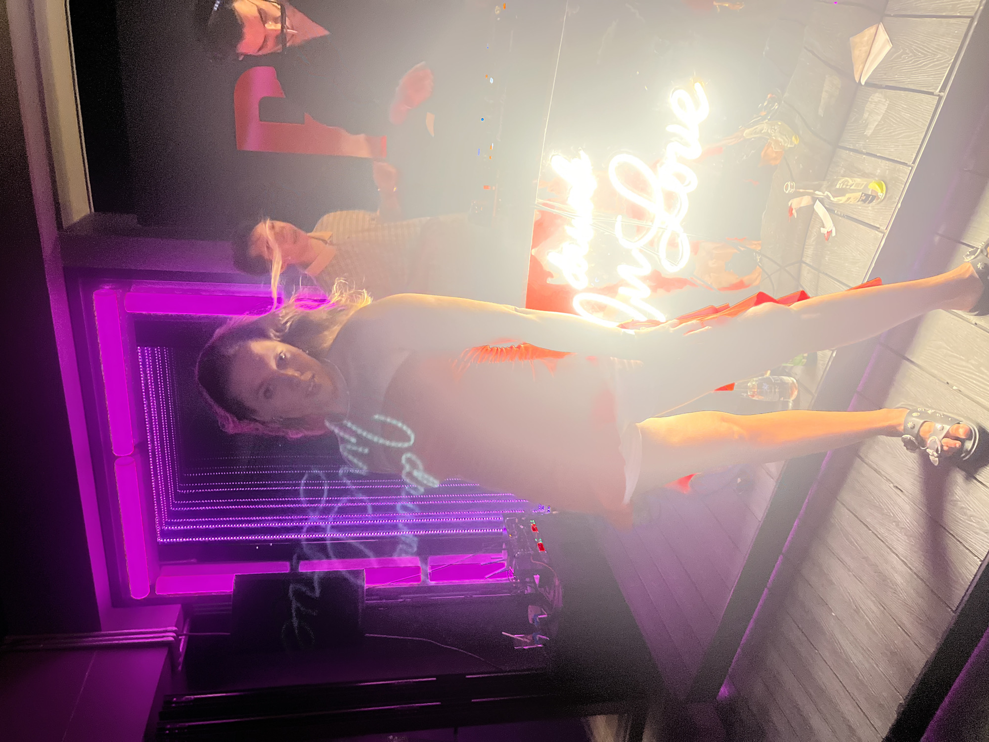 A woman in a strapless red dress and white shorts dances in front of a neon sign that says "Drink in Love". The neon sign is bright white and illuminates the scene with a warm glow. Behind the woman, a DJ booth is visible with a DJ wearing headphones. To the right of the DJ, two men are standing. The environment appears to be an outdoor bar or nightclub in Da Nang, Vietnam, with wooden decking and ambient purple lighting. There are bottles of beer and napkins scattered on the surface in front of the DJ booth. The overall atmosphere suggests a lively night out.
