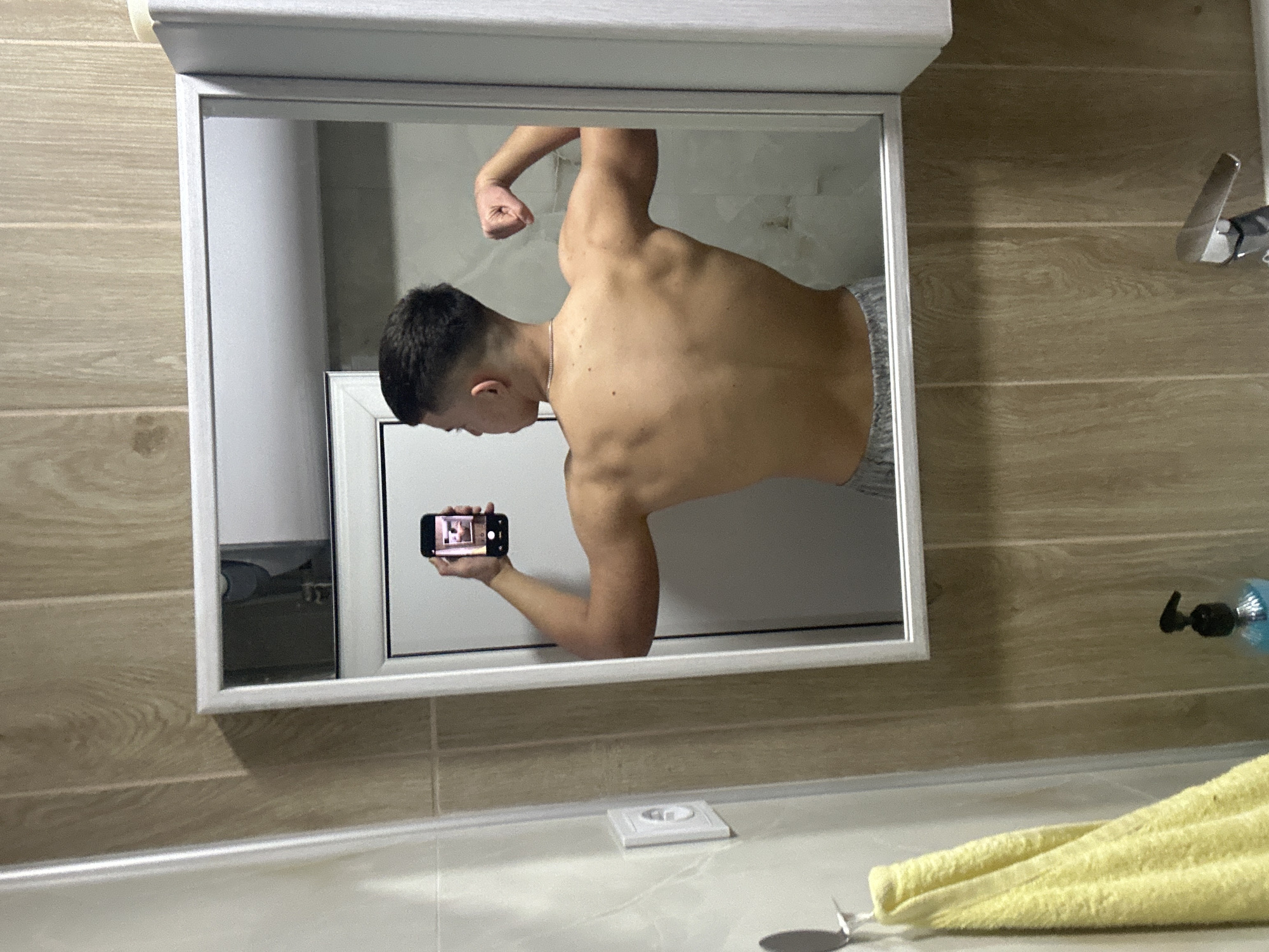 An image captured in a bathroom in Zlatograd, Bulgaria, shows a male individual taking a mirror selfie. The subject is depicted from the back, shirtless, showcasing his muscular physique. He wears light grey athletic shorts or sweatpants. His left arm is flexed, emphasizing the bicep, while his raised right hand holds a black smartphone, reflecting the camera's view of the scene. A thin silver chain is visible around his neck. The bathroom features a rectangular mirror with a white frame, positioned above a white sink equipped with a chrome-finished faucet and a black pump dispenser containing blue liquid. The walls are tiled; one section exhibits light beige-cream tiles, while another features vertical wood-effect tiles. To the left of the mirror, a silver hook supports a bright yellow towel, and a white electrical outlet is present. A white cabinet unit is partially visible to the right of the mirror.