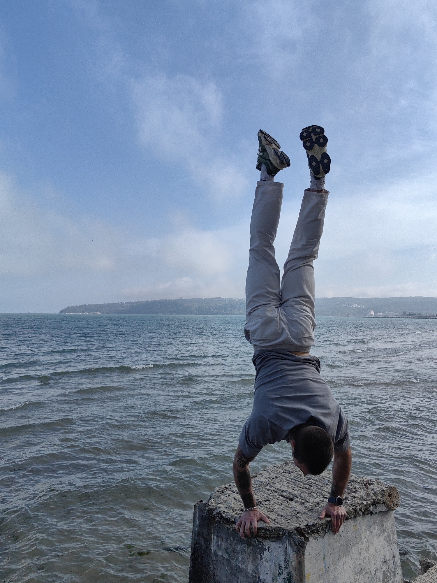 A man is performing a handstand on a concrete structure along the Black Sea coast in Varna, Bulgaria. The man is wearing grey trousers, a grey shirt, and sneakers. He has a dark tattoo on his left arm and a watch on his right wrist. The sea is choppy and stretches into the distance, where the city of Varna is partially visible along the coastline. The sky is partly cloudy.