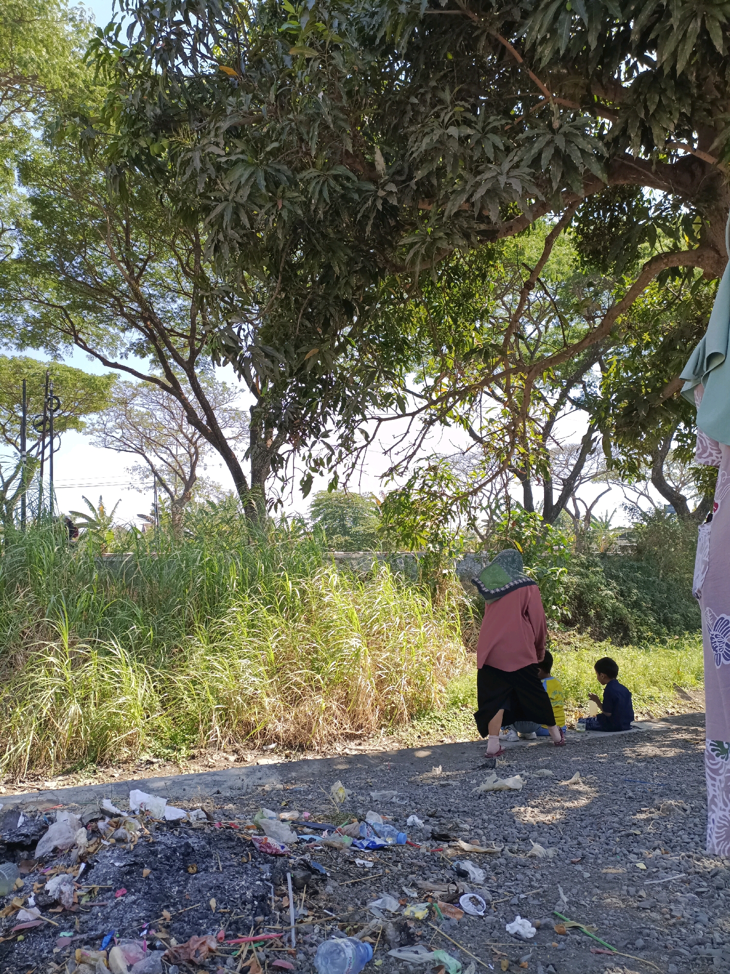 A medium shot shows a roadside scene in Cangkring, Rembang, Indonesia, on a sunny day. In the foreground, a pile of scattered trash, including plastic bags, bottles, and burnt debris, lies on a gravel surface. To the right, a person wearing a light-colored hijab and a maroon top with a black skirt is partially visible, with their back to the camera. They appear to be interacting with a child dressed in a yellow shirt and dark pants, and another child in a blue shirt and dark pants who is sitting on the ground. In the background, tall, lush green grass grows along a gentle incline. Beyond the grass, a variety of trees with dense foliage create a canopy overhead, with some sunlight filtering through. The sky is visible as a pale blue. The scene suggests an outdoor environment, possibly a rural or semi-rural area, with the presence of both natural elements and the evidence of human activity and waste.