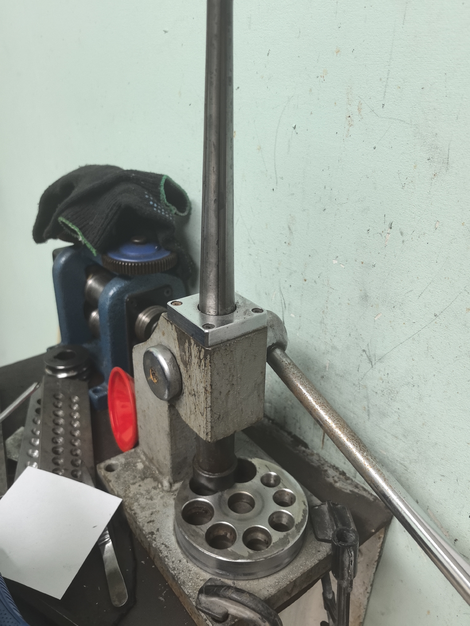 The image shows a close-up of a benchtop metalworking tool, likely a ring-sizing mandrel or a small rolling mill, situated in what appears to be a workshop or industrial setting. The tool is made of metal and has a vertical shaft and a circular base with multiple indentations. To the left of the main tool, there is a blue metal piece with gears and rollers, and to its left, a black work glove is draped over it. There are also various other metal accessories and a small white piece of paper on the workbench. The background is a pale green, textured wall that appears worn and has some marks and scuffs. There is no clear indication of the specific location within Amursk, Russia, but the environment suggests a place of manual labor or craft. There are no people visible in the image.