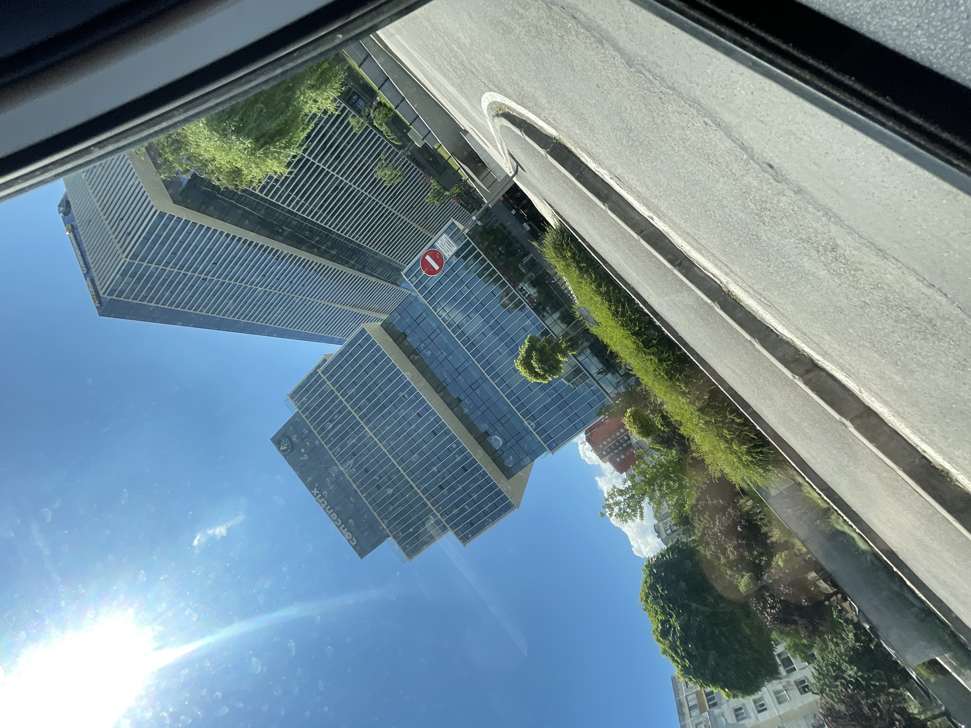 The image is a tilted, wide-angle outdoor shot taken from within a vehicle looking out. The scene depicts a modern office building complex with a prominent sign reading "concentrix" visible on one of the towers. The building has a distinctive stepped architectural design with glass facades. Adjacent to the office complex is a curving road with a traffic sign indicating "No Entry". Lush green trees and bushes are planted along the roadside and around the building. In the background, other buildings, including an apartment block with visible windows, are present. The sky is a clear blue with the sun shining brightly, casting a lens flare. The image appears to be taken during daylight, possibly in the summer. The context of Sofia, Bulgaria is provided, suggesting this is an urban setting within that city.