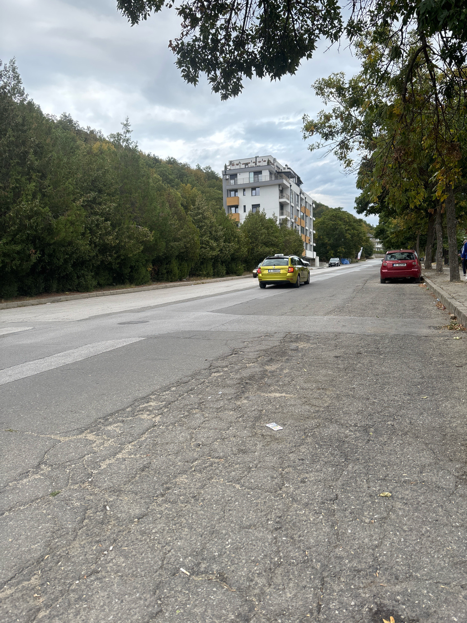 A yellow Skoda Octavia taxi is driving on a road in Blagoevgrad, Bulgaria, on a cloudy day. The taxi is in the left lane of a two-lane road. A red Skoda Fabia is parked on the right shoulder of the road. In the background, there is a white apartment building and a hillside covered in trees. The road is in disrepair, with cracks and potholes visible in the asphalt. There are also some cigarette butts and a piece of paper scattered on the road. A person in blue clothing is walking on the sidewalk to the right of the red Skoda.