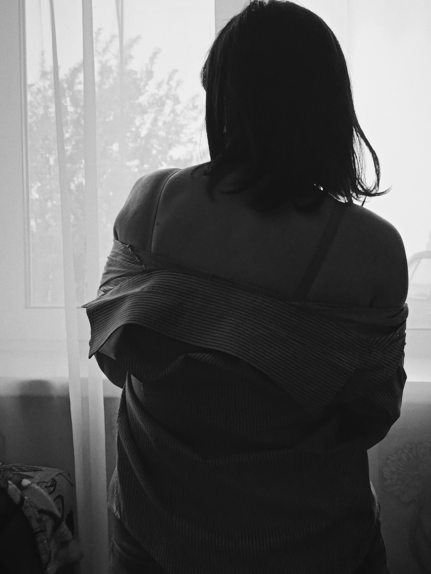 A black and white photograph depicts a woman facing away from the viewer, standing near a window. She has dark hair and is wearing an off-the-shoulder striped shirt. A bra strap is visible on her shoulder. Outside the window, trees are seen, obscured by the curtains. The location is unknown and there is no clear evidence to suggest the photo was taken in Фоки, Russia.