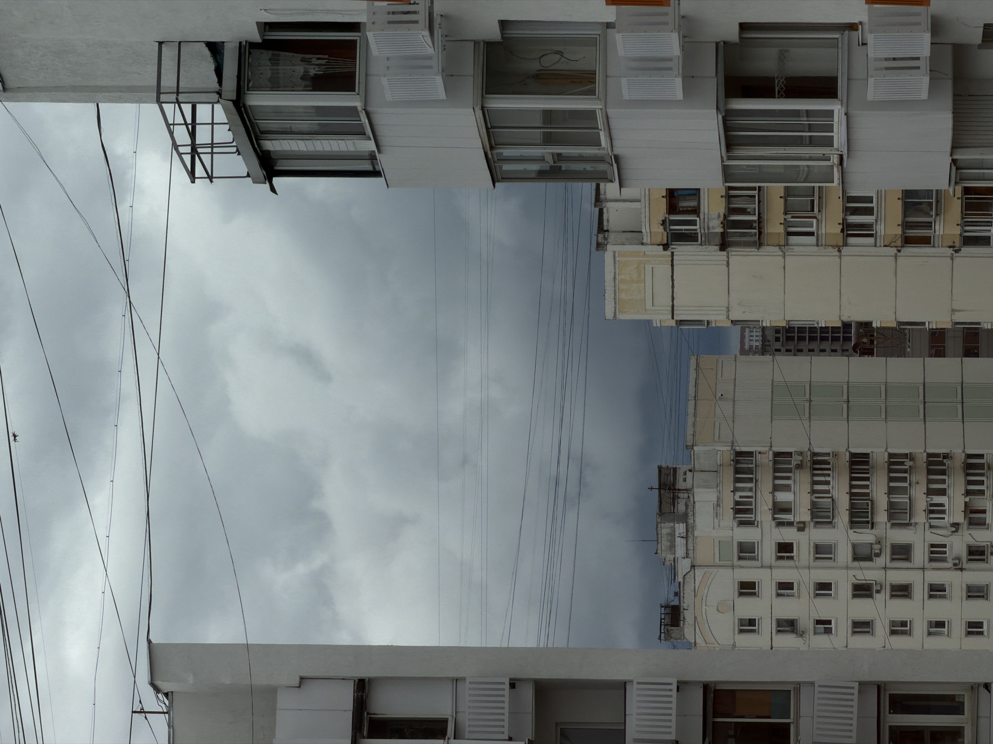 The image shows a view of apartment buildings and a cloudy sky in the city of Khimki, Russia. In the foreground, on the left, is the side of a white apartment building with visible balconies and windows. To the right is another apartment building, also white, with multiple balconies featuring white railings and glass enclosures. Several windows are visible on this building. In the middle ground, several more apartment buildings are visible, with varying architectural styles and colors, primarily beige and white. These buildings have numerous windows and balconies. Overhead, a network of power lines crisscrosses the sky, which is filled with heavy, grey clouds, suggesting overcast or stormy weather. The overall impression is an urban residential scene under a somber sky.