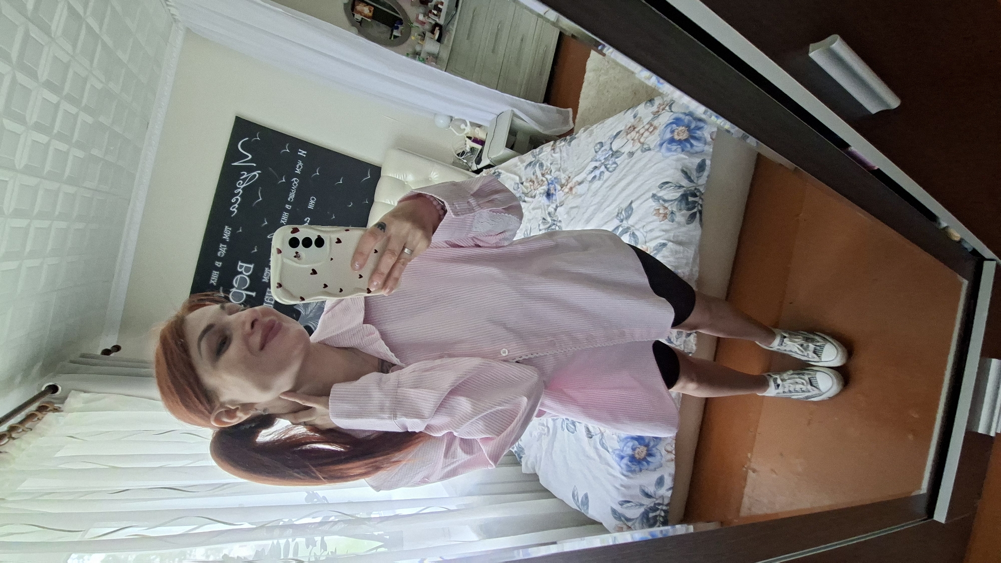 The image shows a young woman with red hair pulled back in a ponytail, taking a selfie in a mirror. She is wearing a light pink, pinstriped oversized button-down shirt and black athletic shorts. Her hair is pulled back in a ponytail. On her hand, she is wearing a ring. In the background, a bed with floral bedding is visible, as well as a dresser with items on top and a large mirror. The setting appears to be a bedroom. There is no clear indication of the city of Myadzyel, Belarus.
