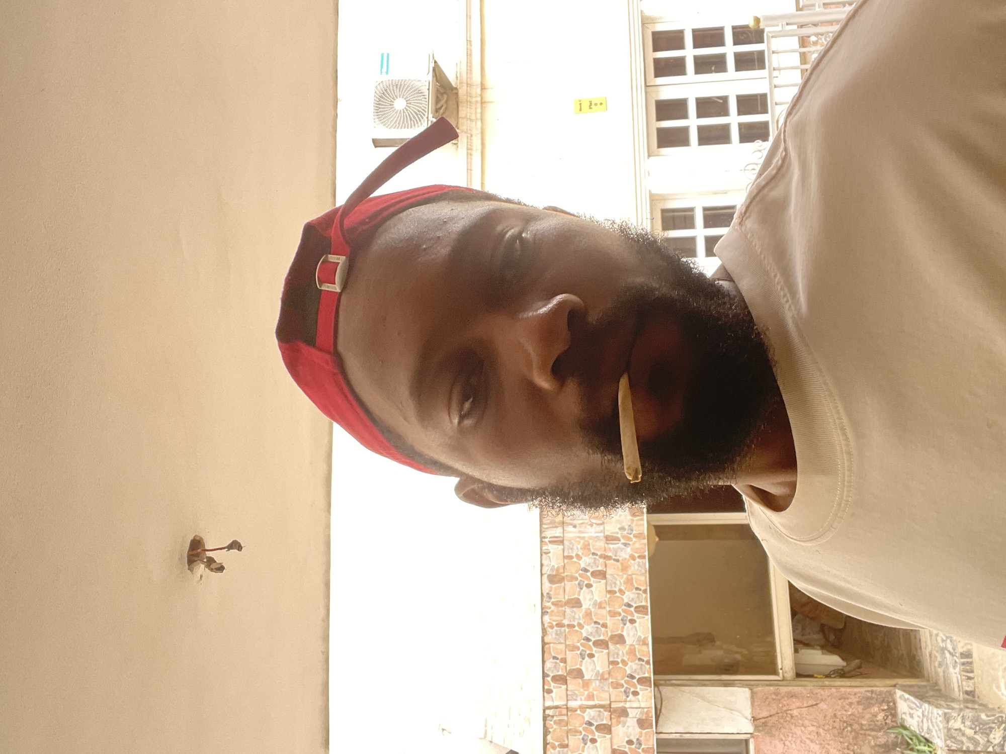 A portrait-style image, rotated 90 degrees clockwise, features a man with dark skin, a full beard, and a red cap worn backward. He is wearing a light-colored (possibly cream or white) t-shirt and has a hand-rolled cigarette, resembling a joint, held between his lips. His eyes are open, and he gazes directly at the viewer with a neutral, calm expression.
The background is divided into several sections. To the man's left, a plain, light-beige wall is visible, featuring a small opening with exposed electrical wires. To his right, there is a wall covered in brown and beige mosaic-like tiles. In the upper right portion of the image, behind the man, a white building with multiple grid-patterned windows is visible, along with an outdoor air conditioning unit mounted on its wall. A small yellow sticker, with symbols that appear to be "1 | 2", is attached near the AC unit. Below the tiled wall, an open window or doorway reveals a glimpse of an interior or another external space.
The scene is brightly lit, suggesting it is daytime. The man is posing for the camera. The architectural style, including the windows, tiled walls, and air conditioning unit, are consistent with an urban or suburban environment, aligning with the provided location context of Abuja, Nigeria.