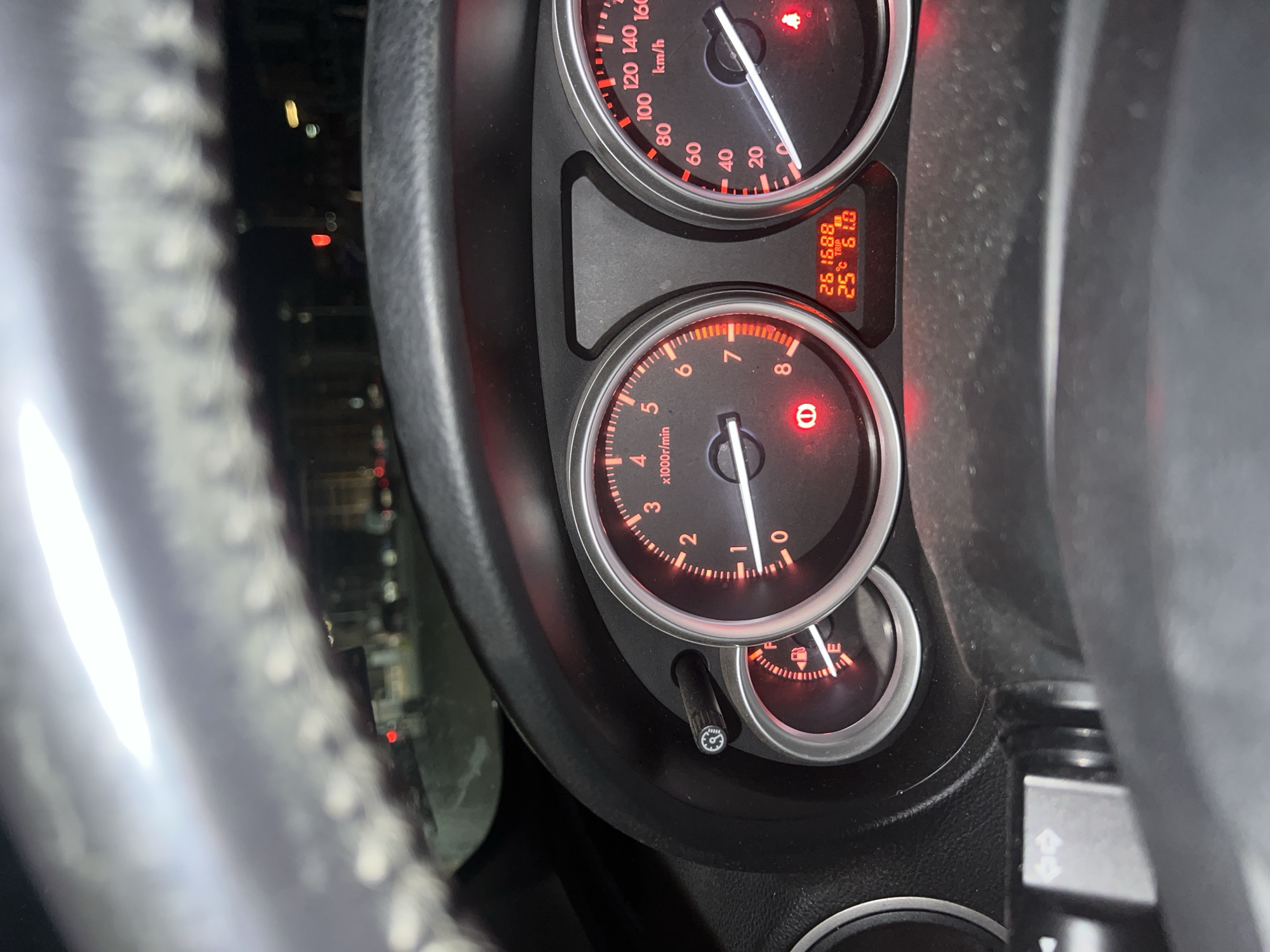 The image depicts a car's dashboard, showing a speedometer reading approximately 25 km/h and a tachometer reading close to 0 rpm. The odometer displays 261688, and the trip meter shows 61.0. The fuel gauge indicates nearly empty. The temperature is displayed as 25 degrees Celsius. The image is taken from the driver's perspective, with a portion of the steering wheel visible on the left. Outside the car window, blurred lights suggest an urban environment at night. There is no indication of the scene being located in Burgas, Bulgaria.