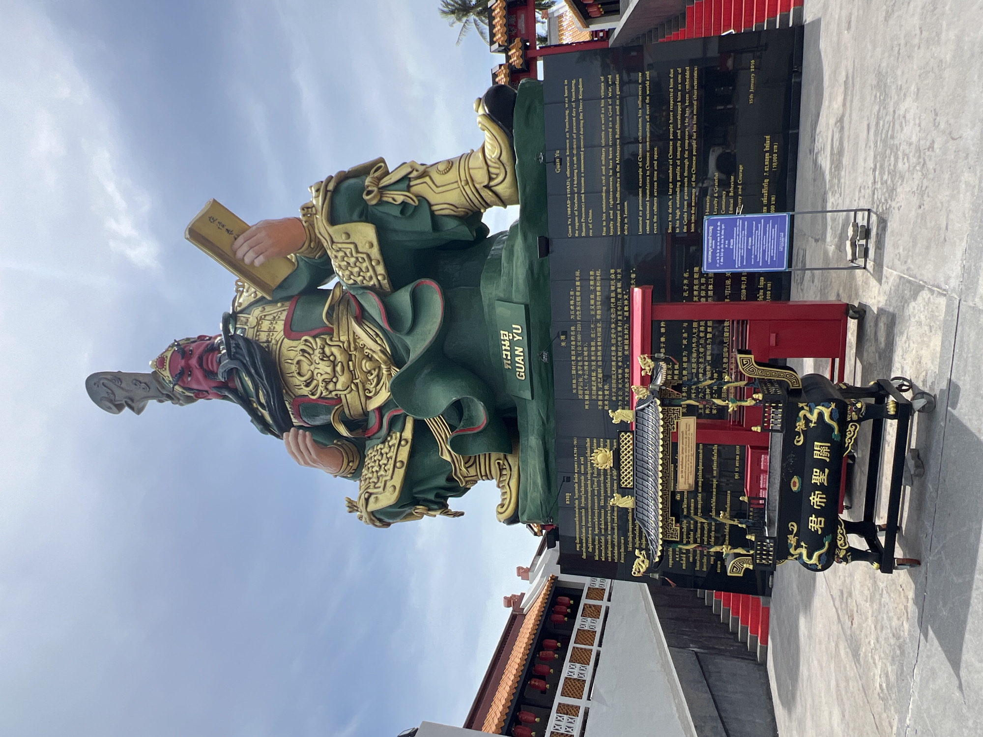 The image depicts a large, seated statue of Guan Yu, a historical figure and deity in Chinese culture, located in Baan Hua Thanon, Thailand. The statue is painted in vibrant colors, with Guan Yu dressed in green armor with gold accents and holding a book. The statue is positioned on a black base adorned with inscriptions in Thai and English, providing information about Guan Yu's life and significance. In front of the statue, there is a black and gold incense burner with Chinese characters and a small red gate. The background features a blue sky with scattered clouds, red-roofed buildings with traditional Chinese architectural elements, and steps leading up to the buildings. The scene appears to be outdoors, likely at a temple or shrine dedicated to Guan Yu.