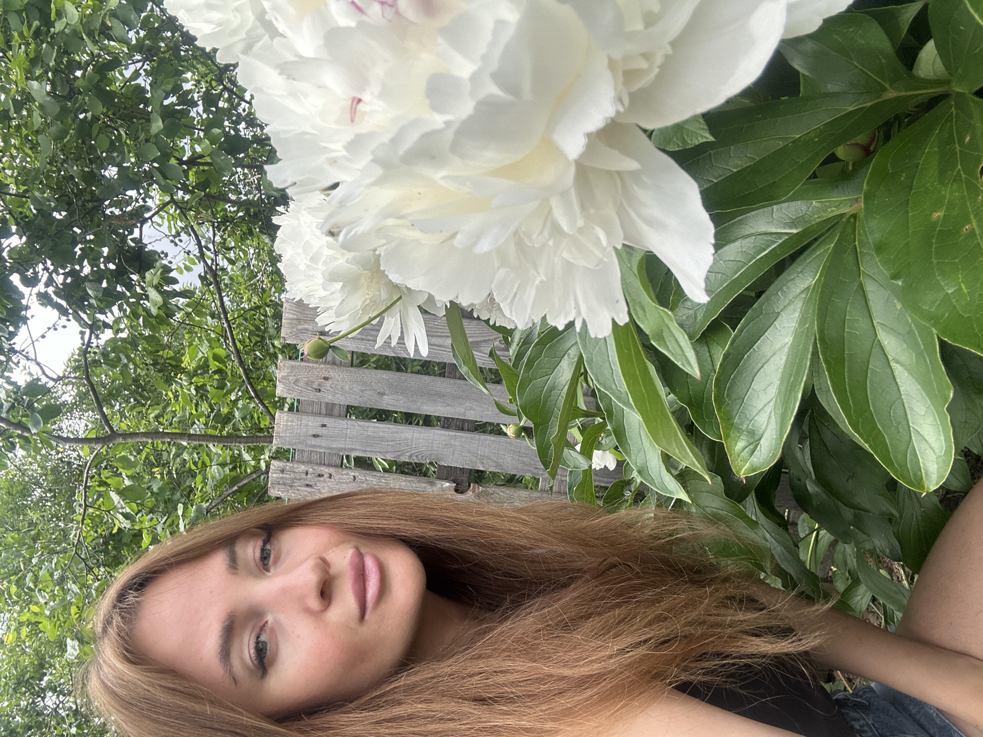 A young woman with long, reddish-brown hair is taking a selfie outdoors. She is looking directly at the camera with a slight smile, her head tilted to the left. She is wearing a black top and denim shorts. In the foreground, a cluster of large white peonies with green leaves is in bloom. Behind her, a weathered wooden fence runs horizontally, and beyond that, lush green trees and foliage suggest a natural, possibly rural or park-like setting. The lighting is natural, indicating it is daytime. The presence of peonies and the weathered fence are common elements found in gardens and yards in Chaikovsky, Russia, a city known for its surrounding natural beauty and recreational areas.