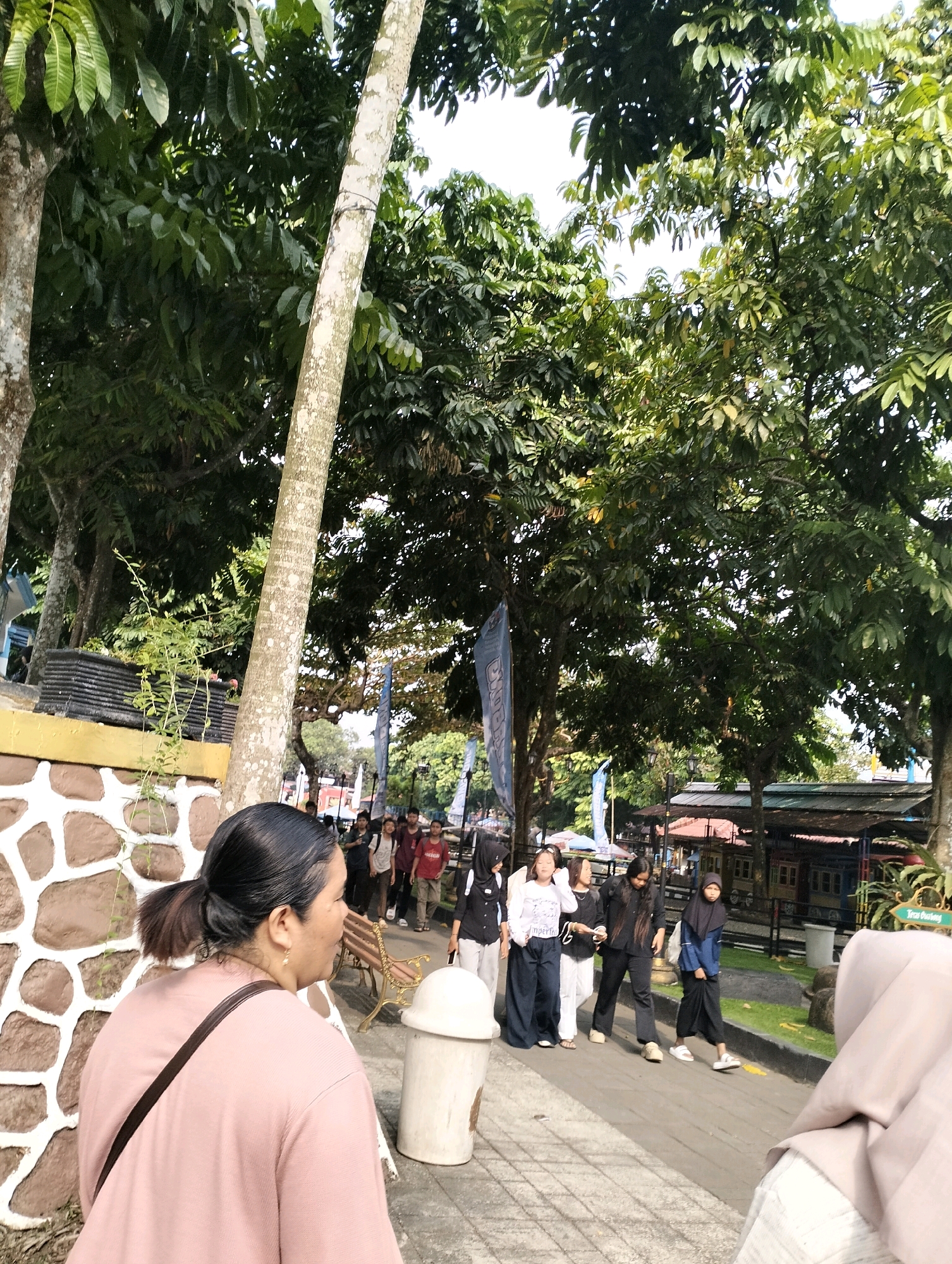 In the city of Karangbanjar, Indonesia, a group of young people walk along a paved path surrounded by lush green trees and foliage. The path is bordered by a low stone wall on the left, constructed with alternating brown and white stones. A tall, slender tree with light-colored bark stands prominently in the foreground, leaning slightly towards the right. In the background, several young individuals, some wearing headscarves and traditional Islamic clothing, walk in the same direction as the viewer. There are also stalls or small buildings visible in the distance, with red and green roofs, suggesting a marketplace or recreational area. A white trash can is positioned near the stone wall. The lighting suggests it is daytime with dappled sunlight filtering through the leaves.