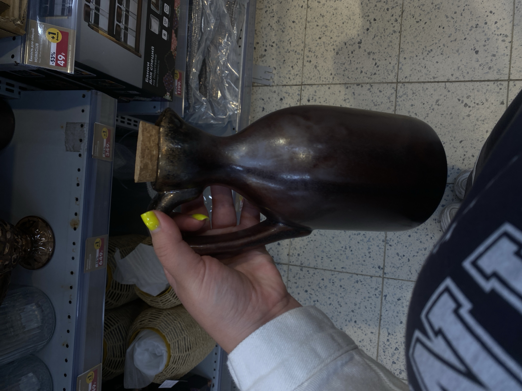 This media file depicts a close-up, low-angle shot of a person's hand holding a dark brown ceramic pitcher with a cork stopper. The pitcher has a smooth, slightly glossy finish and a distinct handle. The hand, with bright yellow nail polish, is positioned to show the pitcher's shape and how it's being held.
In the background, shelves are visible, stocked with various items, suggesting a retail environment. Several labels with Cyrillic text and prices are present on the shelves. One prominent label reads "Емкость для специй HOMECLUB" (Spice container HOMECLUB) with a discounted price of "49P" (49 rubles). Another label indicates "Банки для специй на подставке" (Spice jars on a stand). Some woven baskets are also visible on the shelves. The floor appears to be tiled with a speckled pattern.
The lighting suggests an indoor setting, possibly a store. The overall mood is neutral, focusing on the product and the act of handling it. The text indicates that this is likely in Russia, specifically Peterhof, based on the provided location context. The items on the shelves, including spice containers and baskets, suggest a home goods or kitchenware section of a store.