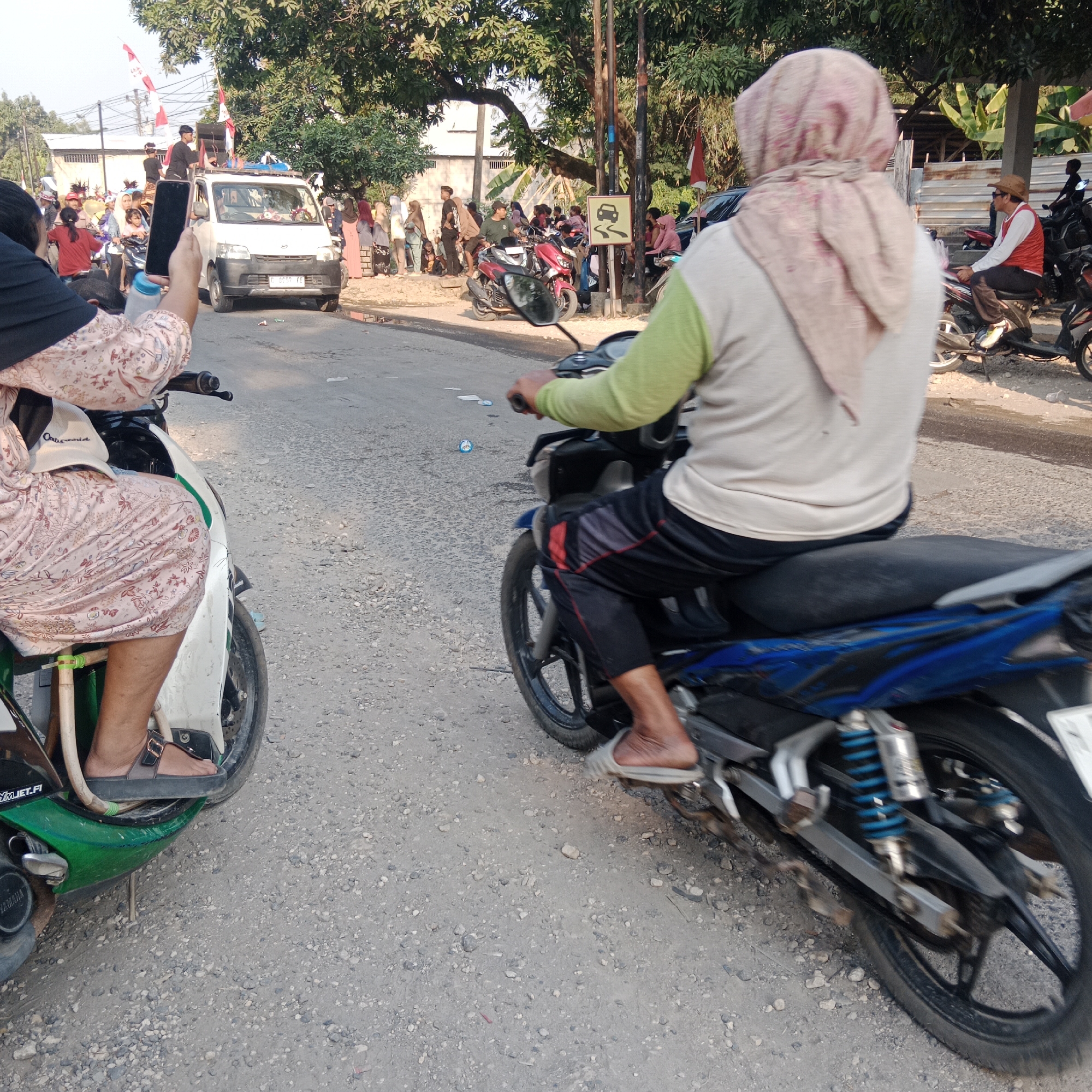 In the city of Tegalrejo, Indonesia, a lively scene unfolds on a gravel road lined with trees. A white van, adorned with Indonesian flags, moves forward, carrying individuals, some of whom are holding speakers. On the side of the road, a crowd of people gathers, watching the procession. The foreground of the image features two motorcycles with riders. One rider, partially visible, is seated on a green and white motorcycle. The other rider, wearing a light-colored long-sleeved shirt and black pants, is on a blue motorcycle. Both riders are looking forward, their actions suggesting they are part of the same event. The overall atmosphere is one of festivity and community engagement.