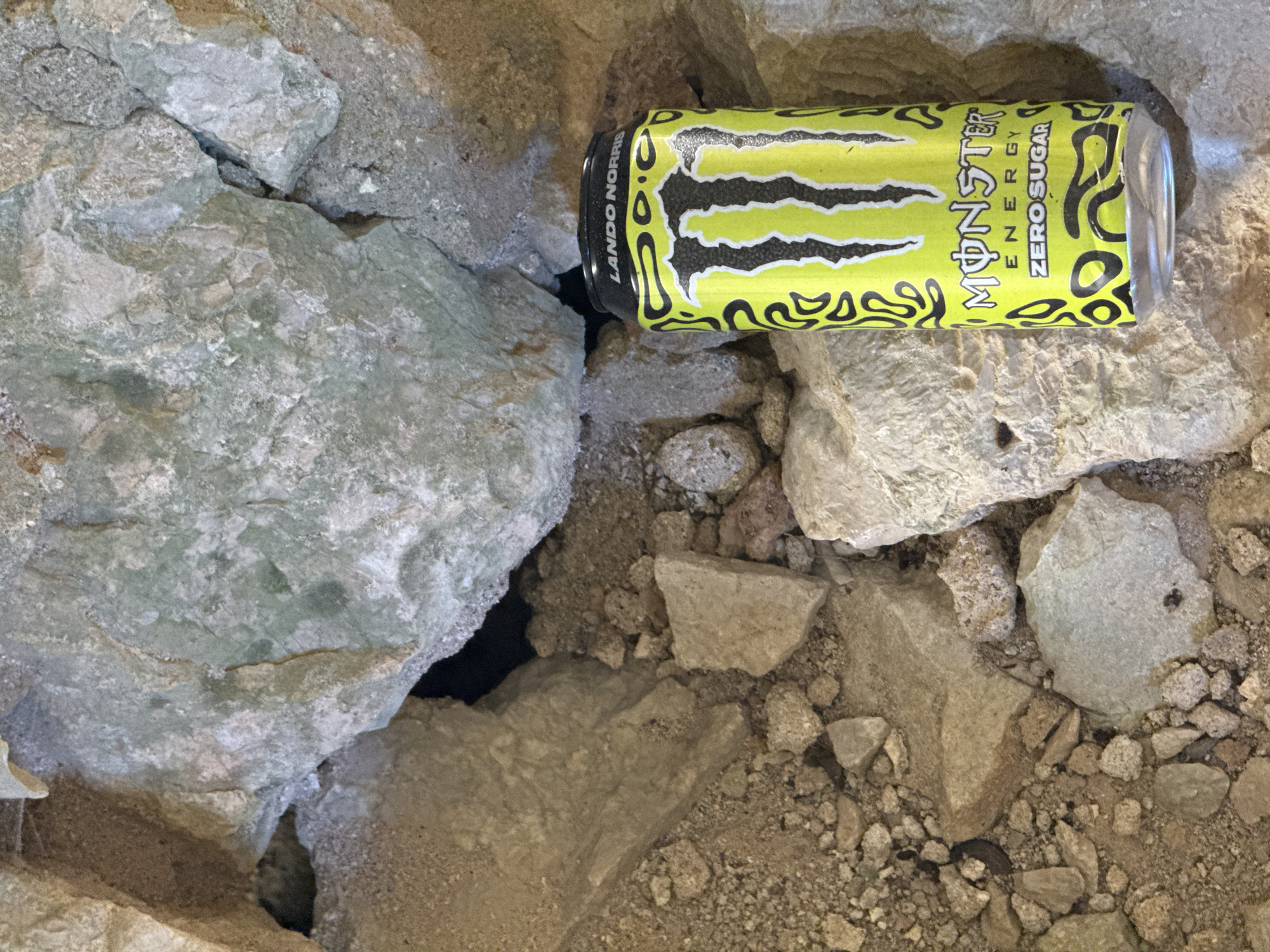 An eye-level, medium shot shows a can of "Monster Energy Zero Sugar" with "LANDO NORRIS" printed on the lid, resting amongst rocks and dirt in Brajici, Montenegro. The can is positioned vertically in the lower right quadrant of the image. The surrounding environment consists of rough, greyish-green and tan rocks, some with a coarse texture, and loose soil and gravel. The background features a dark opening or crevice between the rocks. The lighting appears to be natural, casting soft shadows. There are no people or visible actions taking place in the scene.
