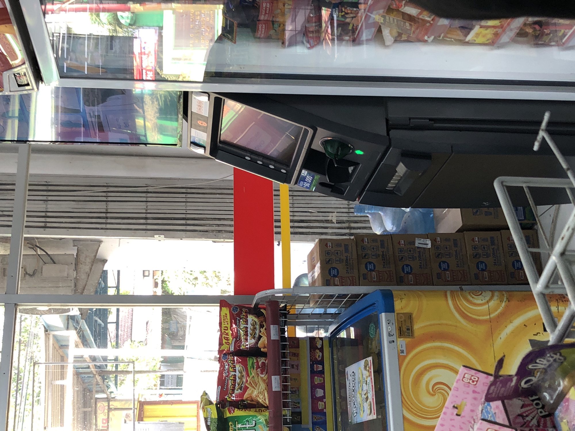 This image shows an interior view of a minimarket in Putat, Indonesia. In the foreground, a gray ATM machine is visible, displaying a green light indicating it is operational. To the right of the ATM, a glass-fronted display refrigerator is filled with packaged snacks. Behind the ATM, stacked cardboard boxes, possibly containing beverages, are visible. To the left of the ATM, shelves hold various packaged food items, including bags of "French Fries" potato chips. A large, yellow and orange swirl-patterned freezer is also present on the left side of the image. In the background, through a large window, an outdoor area with structures and vegetation can be seen. The scene suggests a typical retail environment. No people are clearly visible in the frame.