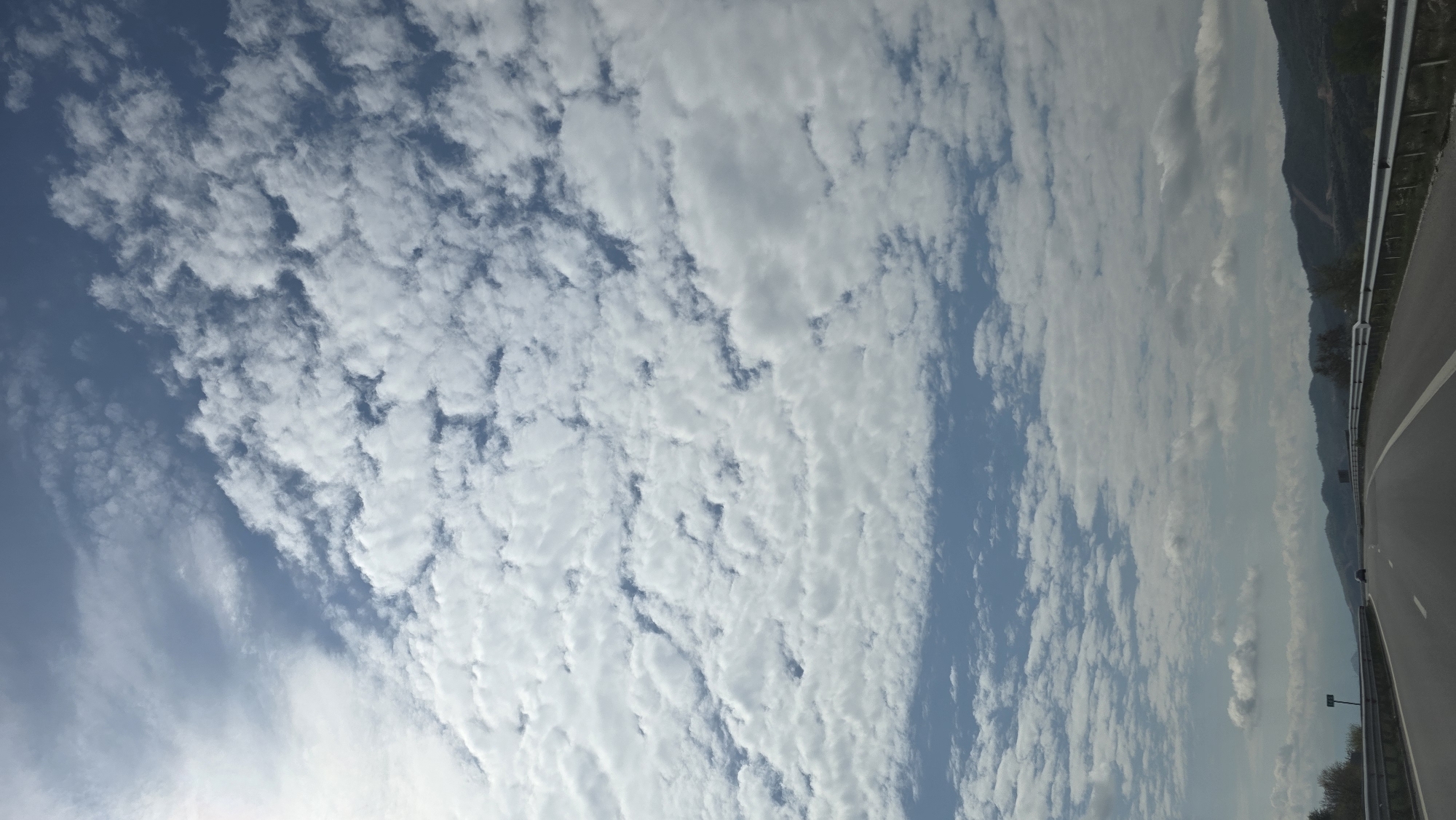 The image shows a daytime sky filled with altocumulus clouds above a highway in Valkovo, Bulgaria. The clouds exhibit a rippled, layered pattern. The highway, visible on the right side of the image, is bordered by a metal guardrail and possibly features mountainous terrain in the background.