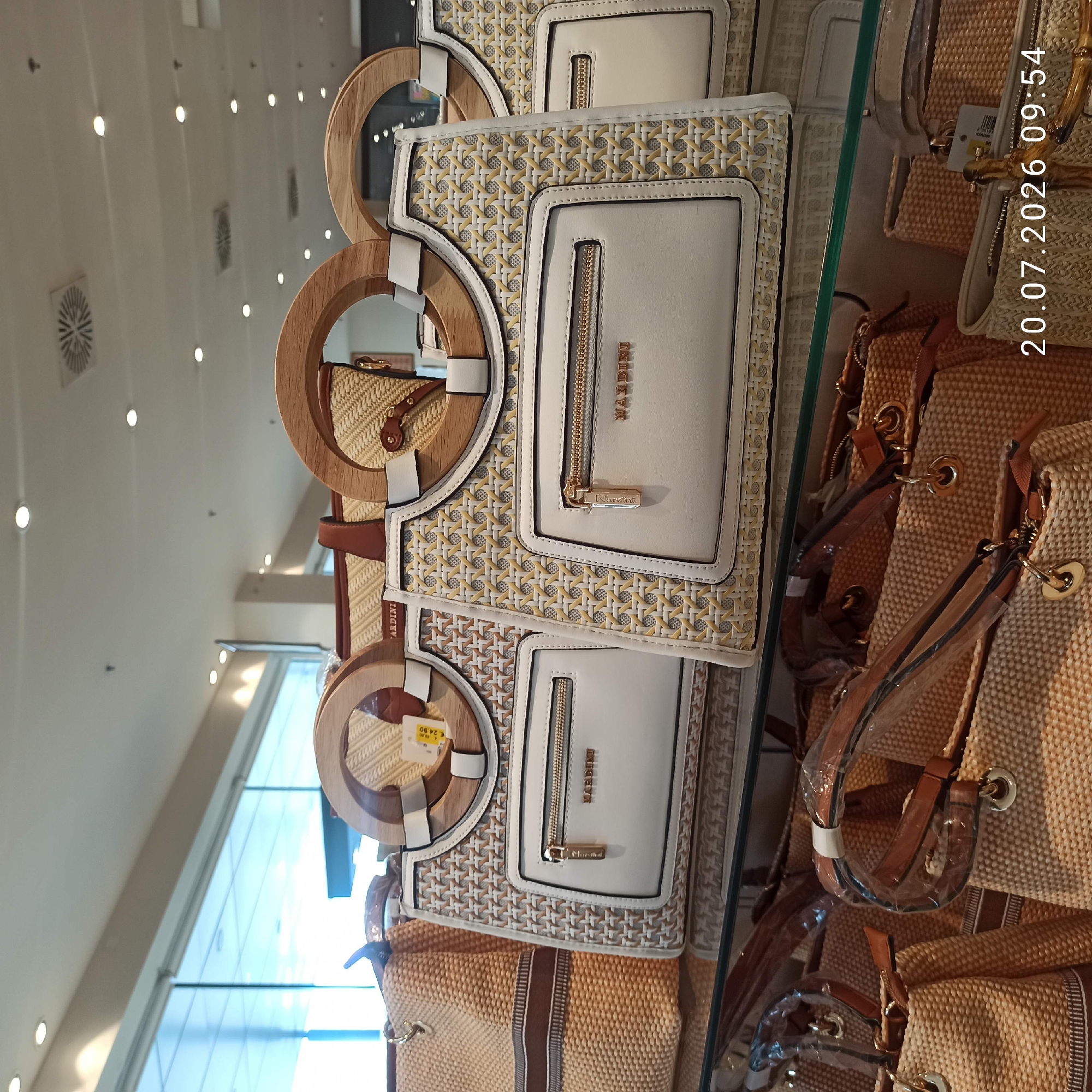The image displays a retail store interior, likely a department store or boutique, with a focus on handbags. The handbags are presented on glass shelves. In the foreground, two white and woven handbags with distinctive wooden hoop handles are prominently displayed. These bags feature multiple zippered compartments and branding that appears to read "MARDINI." To the left of these, a larger woven straw-colored bag with brown trim is visible. Below these, a collection of similar woven straw bags, some with plastic protective wrapping, are arranged on a lower shelf. The background reveals a bright, modern retail environment with large windows, ceiling lights, and ventilation systems, suggesting a commercial setting. The timestamp in the lower right corner reads "20.07.2026 09:54." The scene does not depict any people.