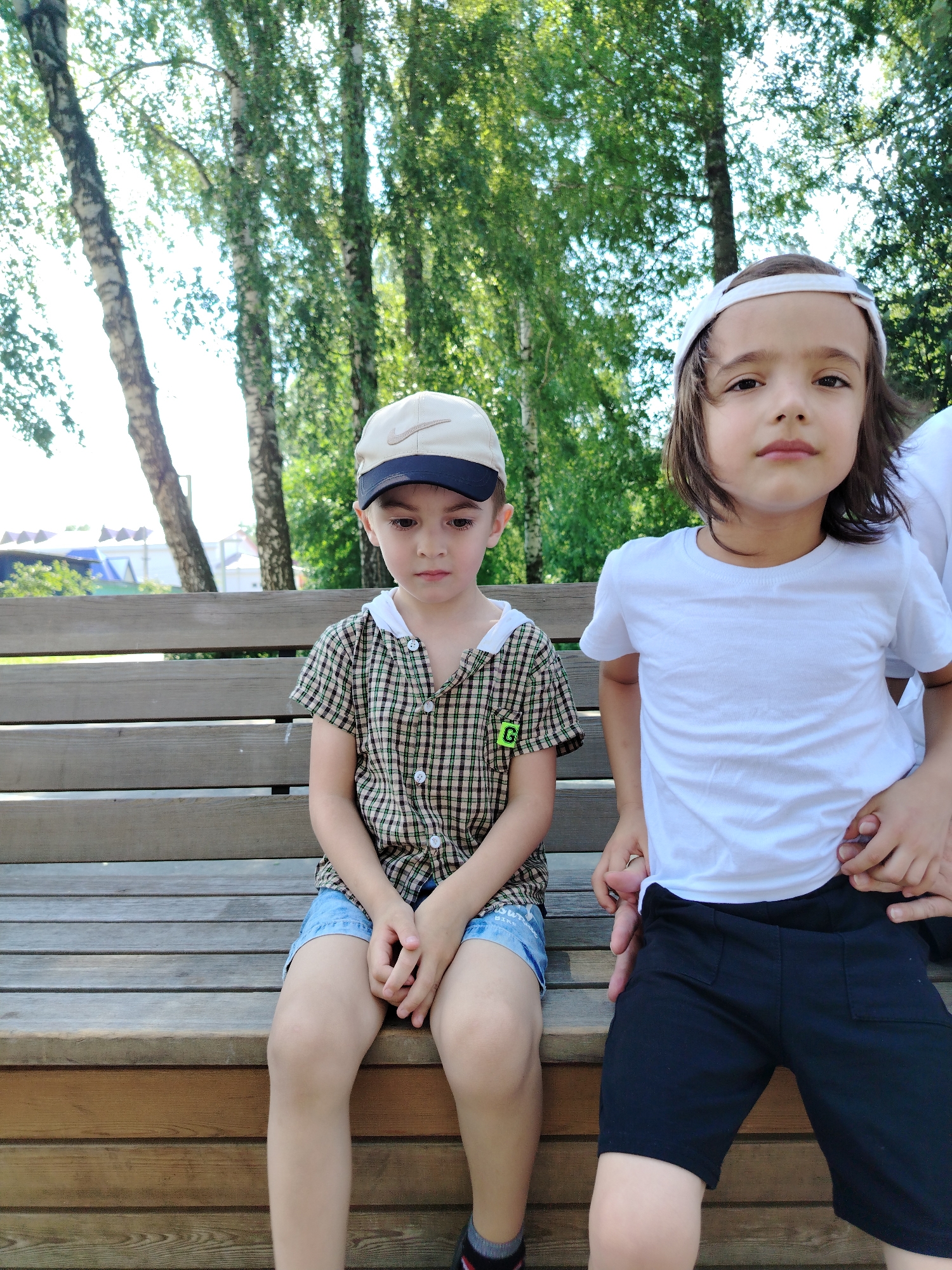 Two young boys are seated on a wooden bench outdoors, with a backdrop of birch trees and foliage. The boy on the left is wearing a plaid collared shirt, denim shorts, and a tan baseball cap with a dark brim and a Nike logo. He is looking down with his hands clasped. The boy on the right, wearing a white t-shirt and black shorts, is looking towards the camera. His right hand is resting on his lower abdomen, with another hand on top of it. The setting appears to be a park or recreational area in Yanaul, Russia. The lighting suggests it is daytime, with sunlight filtering through the leaves.