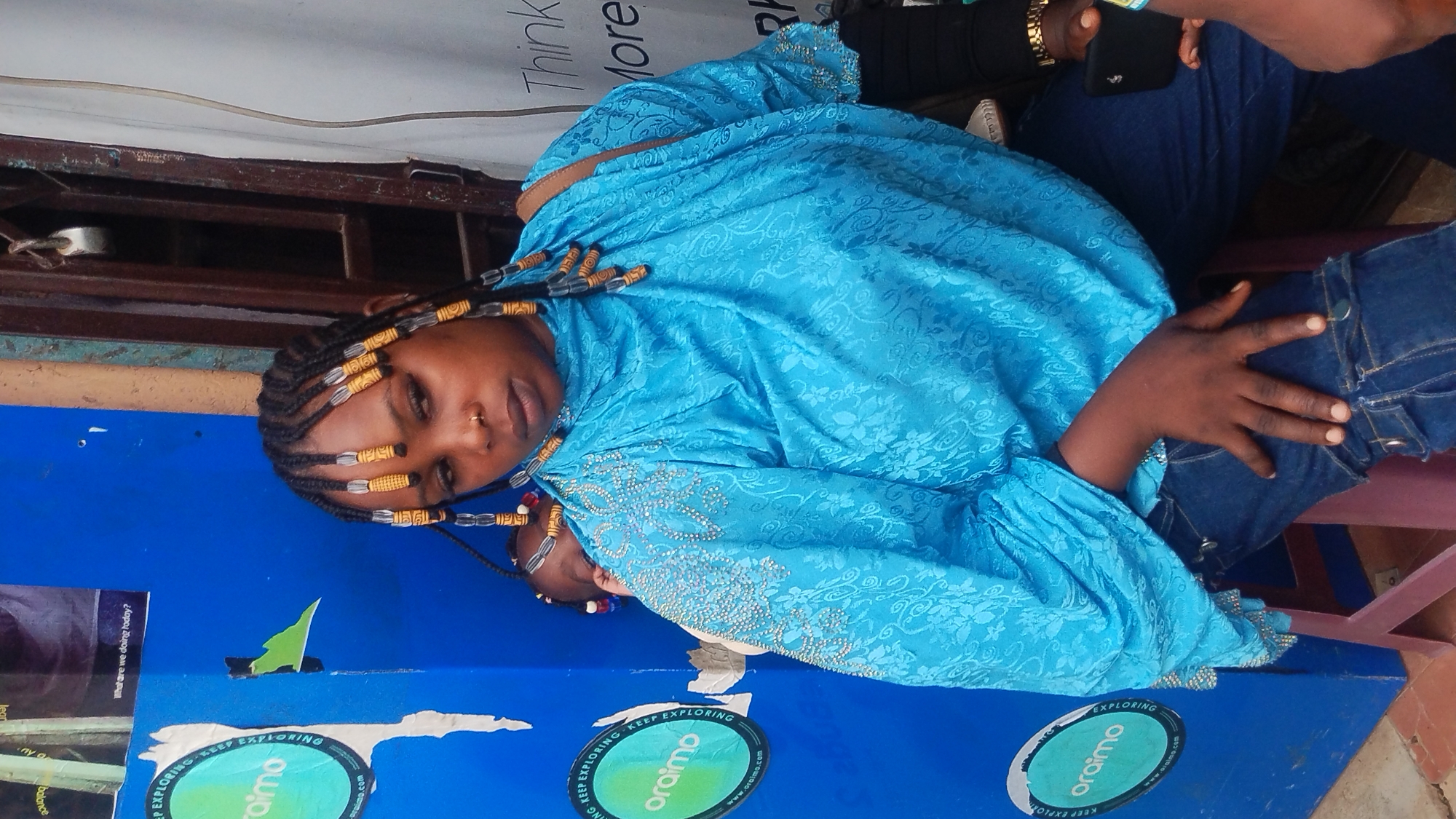 This image, rotated for proper viewing, captures a young person, likely a girl, of color looking directly at the camera with a calm expression. She is wearing a striking bright blue, long-sleeved garment adorned with intricate floral patterns and sparkling embellishments, suggesting traditional or festive attire. Her dark hair is styled in numerous braids, each decorated with cylindrical yellow and silver beads. A small nose ring is visible on her face.
She appears to be seated or leaning against a prominent blue surface in the foreground, which is covered with several round stickers. Part of a smaller child, indicated by a hand and beaded hair, is partially visible beneath her left arm. To her right, another person's arm, wearing a watch, holds a black smartphone, with their leg in denim jeans also visible. The setting appears casual, possibly an indoor or semi-outdoor space.
The blue surface in the foreground displays multiple stickers with the text "KEEP EXPLORING" and "oraimo" along with "oraimo.com." Other partial text visible on posters or signs includes "balance," "nyo," "What are we doing today?", and "Think More" in the upper background. The background also features a weathered brown structure, possibly a shelf or window frame, adding to the informal ambiance. The lighting suggests it is daytime.