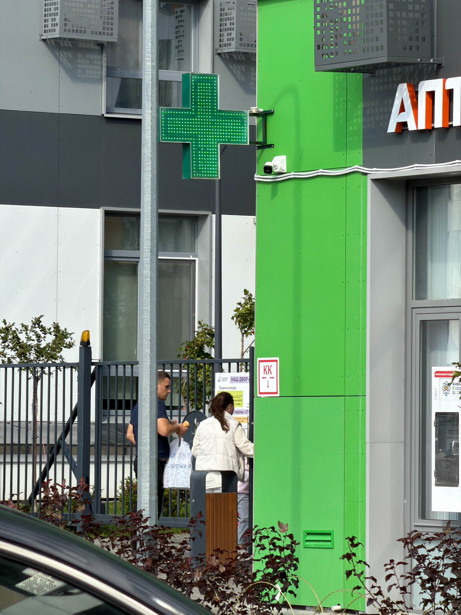 In an outdoor urban setting, a bright green pharmacy cross sign is prominently displayed on the facade of a modern building. The building features a mix of grey and white paneling, with some windows visible. To the right of the pharmacy sign, the white and orange letters "АПП" are visible, suggesting the name of the establishment. A security camera is mounted on the green facade below the cross.
In front of the building, a metal fence encloses a small area with some greenery. Behind the fence, a man in a dark blue shirt is holding a white plastic bag, and a woman in a light-colored jacket is standing near an entrance. There is a small sign with red lettering and arrows, indicating directions. The ground in the foreground is partially occupied by a dark-colored car. The overall scene suggests a commercial area, possibly a street or parking lot in front of a pharmacy or medical facility.