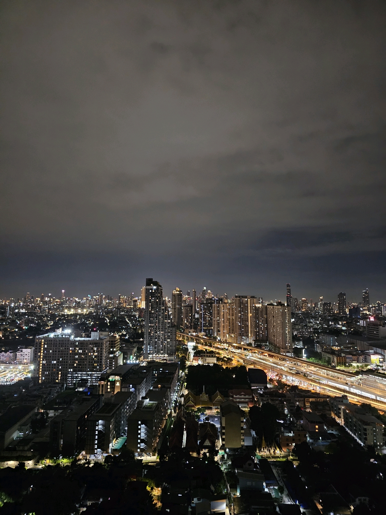 The image shows an aerial view of Bangkok, Thailand at night. The cityscape is dominated by tall buildings, illuminated by numerous lights. A complex network of elevated roads and train tracks is visible, with streaks of light indicating moving vehicles. In the foreground, there are several residential buildings, some with lit windows and a swimming pool. The sky is overcast with dark clouds.