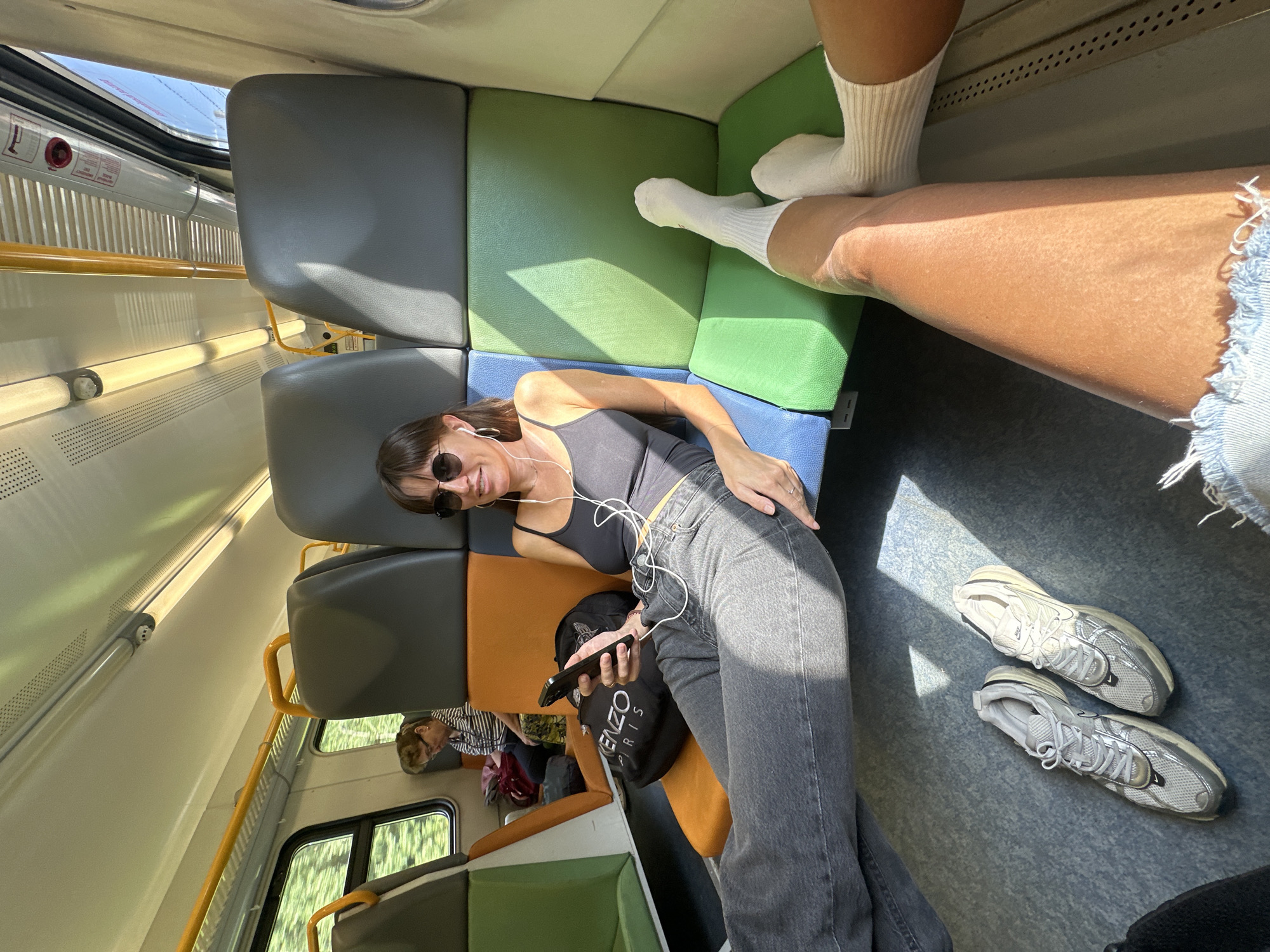 In a train carriage at Stantsiya Petelino, Russia, two individuals are seated. In the foreground, a woman with sunglasses and earbuds is looking at her phone, dressed in a black tank top and grey jeans. Her feet, clad in white socks, are extended onto the seat in front of her. To her left, a pair of grey sneakers rests on the train floor. Further down the aisle, another woman is seated, wearing a striped shirt and glasses, with a red bag beside her. The train seats are multi-colored, featuring orange, blue, and green sections. Sunlight streams through the windows, casting shadows across the interior.