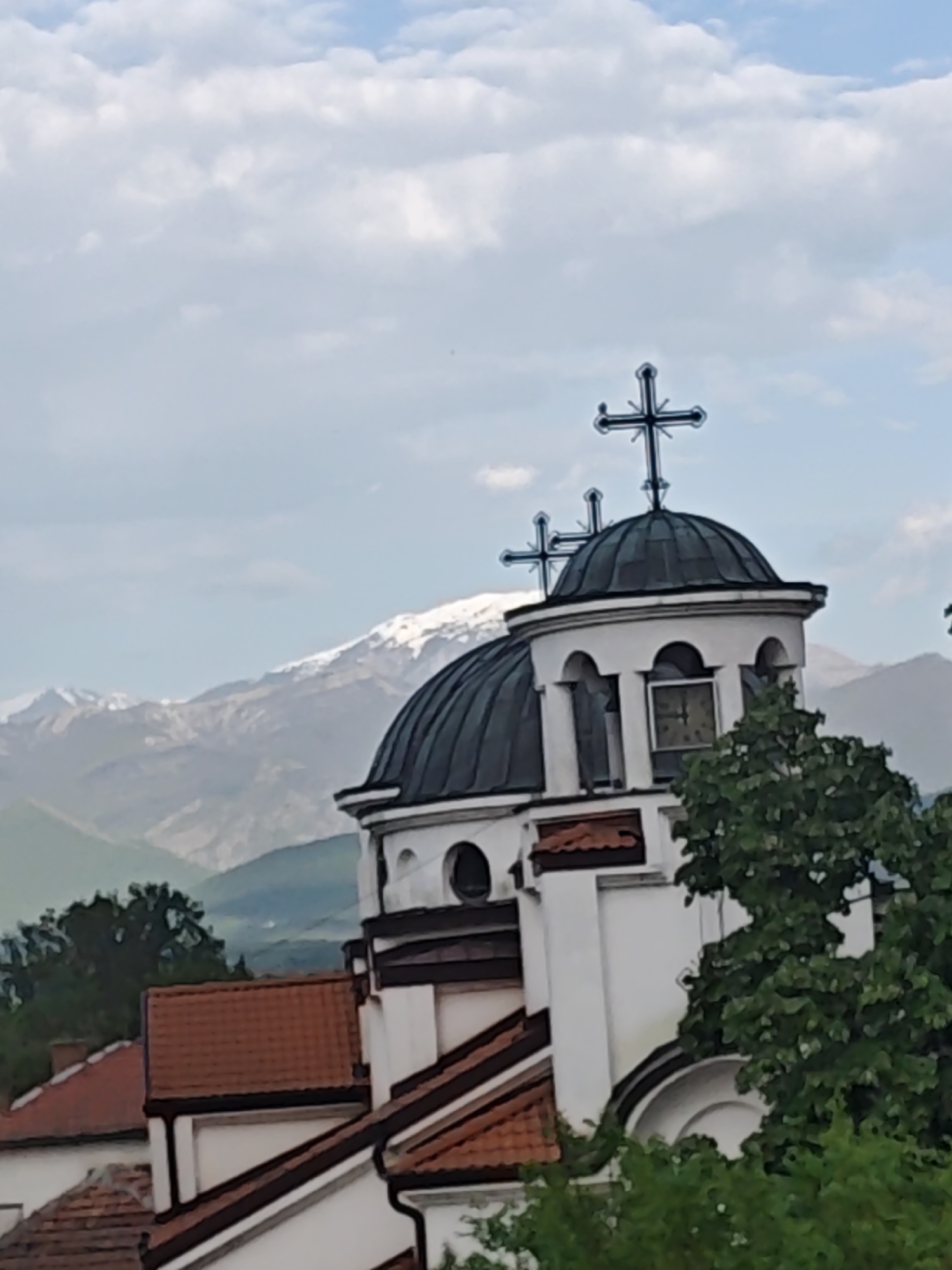 An outdoor, eye-level shot captures a white Orthodox Christian church with dark domes and crosses, set against a backdrop of snow-capped mountains under a cloudy sky. The main focus is the upper section of the church, featuring a large central dark dome topped with an ornate cross, and a smaller turret-like structure to its right, which also has a dome and a cross, along with a visible clock face displaying Roman numerals. The church walls are white, complemented by dark trim and reddish-brown tiled roofs on its lower sections.
In the mid-ground, vibrant green trees partially frame the church on the right and lower left. Beyond the church, a range of majestic mountains rises, with their peaks distinctly covered in snow, suggesting either high altitude or a cooler season. The sky above is mostly filled with soft, white clouds, with hints of blue peaking through. Partial views of other buildings with terracotta-tiled roofs are visible in the lower left corner, indicating a settlement or village surrounding the church. The lighting suggests it is daytime, possibly late morning or early afternoon, with a serene and calm atmosphere. No specific activity is taking place, and the image is a static architectural and landscape view. The numbers on the clock face are not clearly legible.