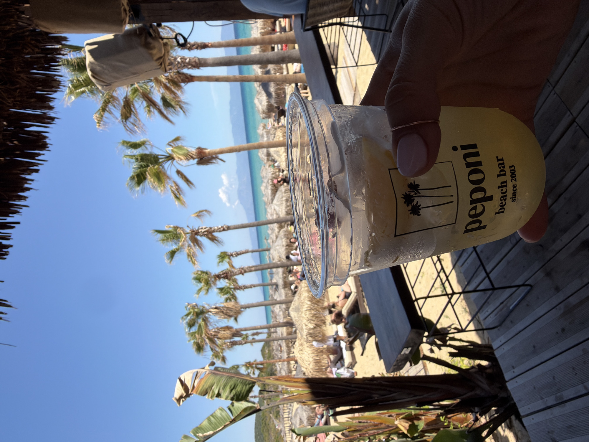 The image is taken from a low-angle perspective at Peponi beach bar in Nea Peramos, Greece. The scene captures a hand holding a plastic cup filled with a yellow drink, presumably a cocktail, adorned with condensation and ice. The cup features the "Peponi beach bar since 2003" logo with a palm tree design. In the background, the beach stretches out with several thatched umbrellas and sunbathers dotting the sand. Palm trees line the beach, leading to a serene view of the blue sea meeting the sky. The angle also includes parts of the beach bar's wooden deck and a glimpse of the bar's structure. The weather appears sunny and clear.