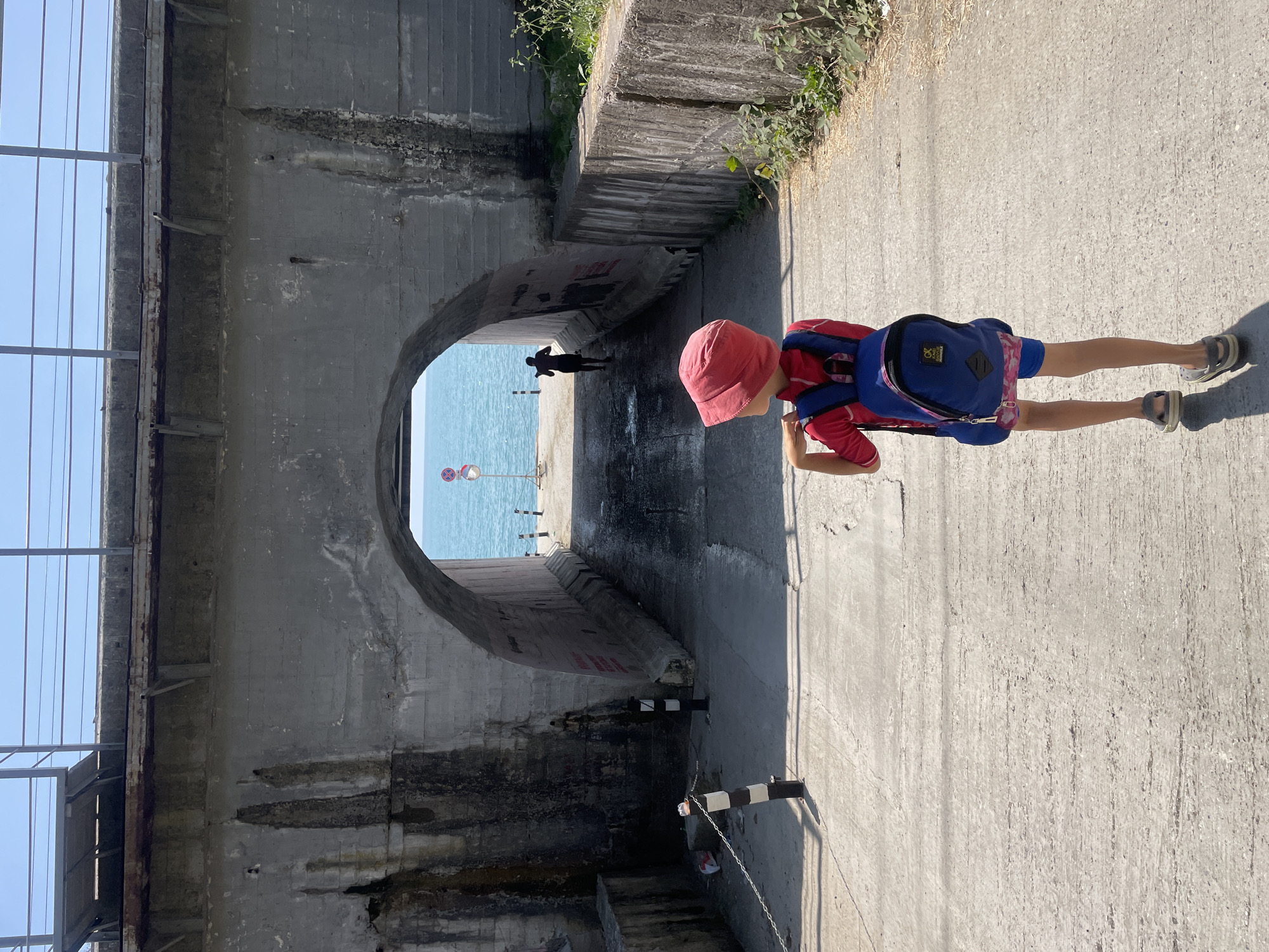 A young child wearing a red hat, red shirt, and blue backpack walks away from the camera on a concrete path towards a large concrete archway. The archway opens to a view of the bright blue sea and sky. Another person, silhouetted and wearing dark shorts, stands facing the sea within the archway. The scene appears to be in Sochi, Russia, given the context of the location. The environment is coastal, with rough concrete structures and some sparse vegetation.