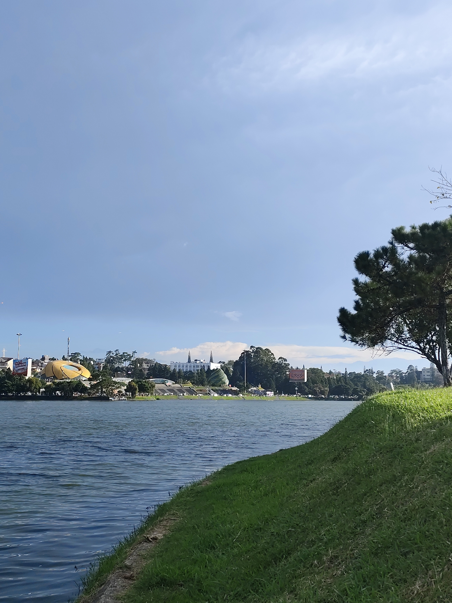 The image depicts a serene landscape view of Xuân Hương Lake in Đà Lạt, Vietnam. The foreground is dominated by a grassy slope leading down to the water's edge. The lake itself is calm, reflecting the blue sky and the cityscape on the opposite bank.
The cityscape features a mix of architectural styles, with prominent buildings including a distinctive yellow, petal-shaped structure, and other structures with white facades and pitched roofs, some resembling churches. Trees and greenery are interspersed throughout the urban skyline, softening the transition between the city and the natural environment. The sky is a clear blue with some wispy clouds, suggesting fair weather. The overall impression is one of a peaceful urban park setting, with the lake serving as a central focal point. There are no visible people or animals in the frame.