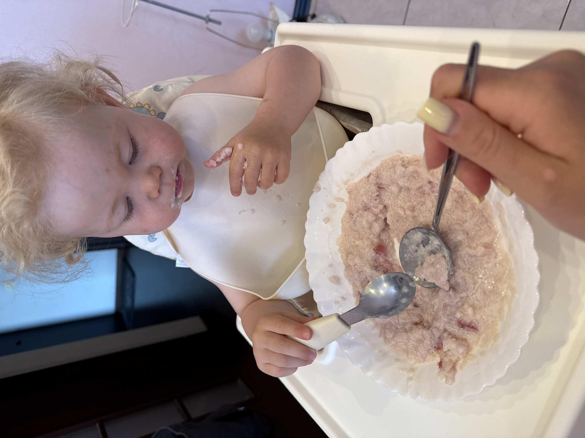 In a high chair, a young child with blond hair wearing a white bib is being fed. The child's face and hands are smudged with food, indicating a messy mealtime. A bowl of pale pink food, likely pureed or mashed, is in front of the child. Two spoons are visible: one held by the child, and another held by an adult whose hand with light yellow fingernails is in the foreground, offering food to the child. The background is blurred but suggests an indoor setting with furniture and a floor. The location is not discernable from the image.