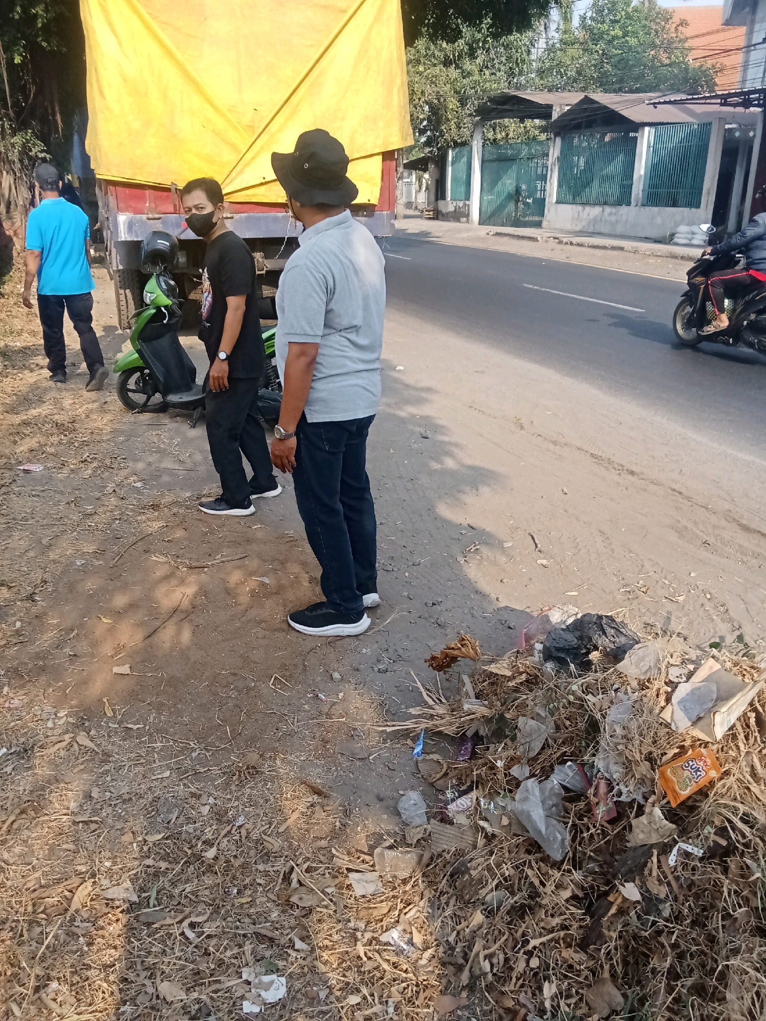 In Kaliwungu, Indonesia, on a roadside with a dirt and gravel shoulder, a yellow tarp-covered truck is parked. Three individuals are present near the truck. One man, wearing a black hat and gray shirt, stands with his back to the camera, facing away from the truck. Another man, in a black t-shirt and black pants, stands to the left of the first man, also facing away from the truck. A third person, wearing a blue t-shirt and dark pants, is further left, walking away from the truck. A green motorcycle is parked behind the man in the black t-shirt. A pile of trash, including plastic bags, dried leaves, and a food wrapper, is visible on the roadside to the right of the men. A road with a motorcyclist in the distance is also visible. The environment appears to be a sunny day.