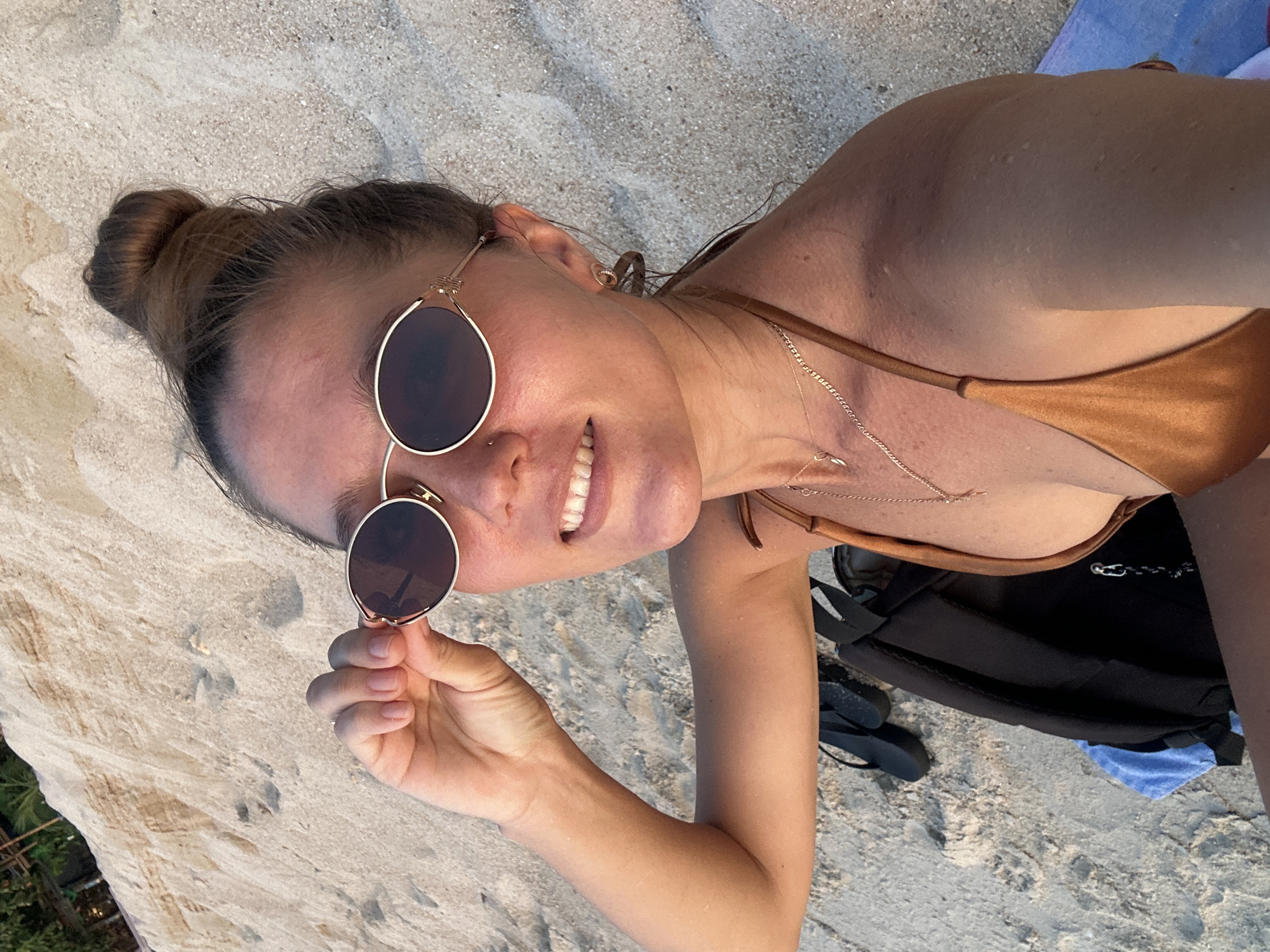 A young woman with her hair in a bun is lying on a sandy beach, taking a selfie. She is wearing a bronze bikini and round, rose gold sunglasses. The woman is smiling, with her teeth visible, and appears relaxed. A thin necklace adorns her neck. Beside her, on the sand, is a black backpack and a pair of flip-flops. The scene is set in a tropical location, suggested by the sandy beach and what appears to be lush greenery in the lower left corner of the frame. The lighting indicates it is daytime, likely sunny, given the shadows and the bright exposure. The overall mood is casual and leisurely, consistent with a beach vacation. There is no visible text in the image. The location is identified as Ko Samui District, Thailand.