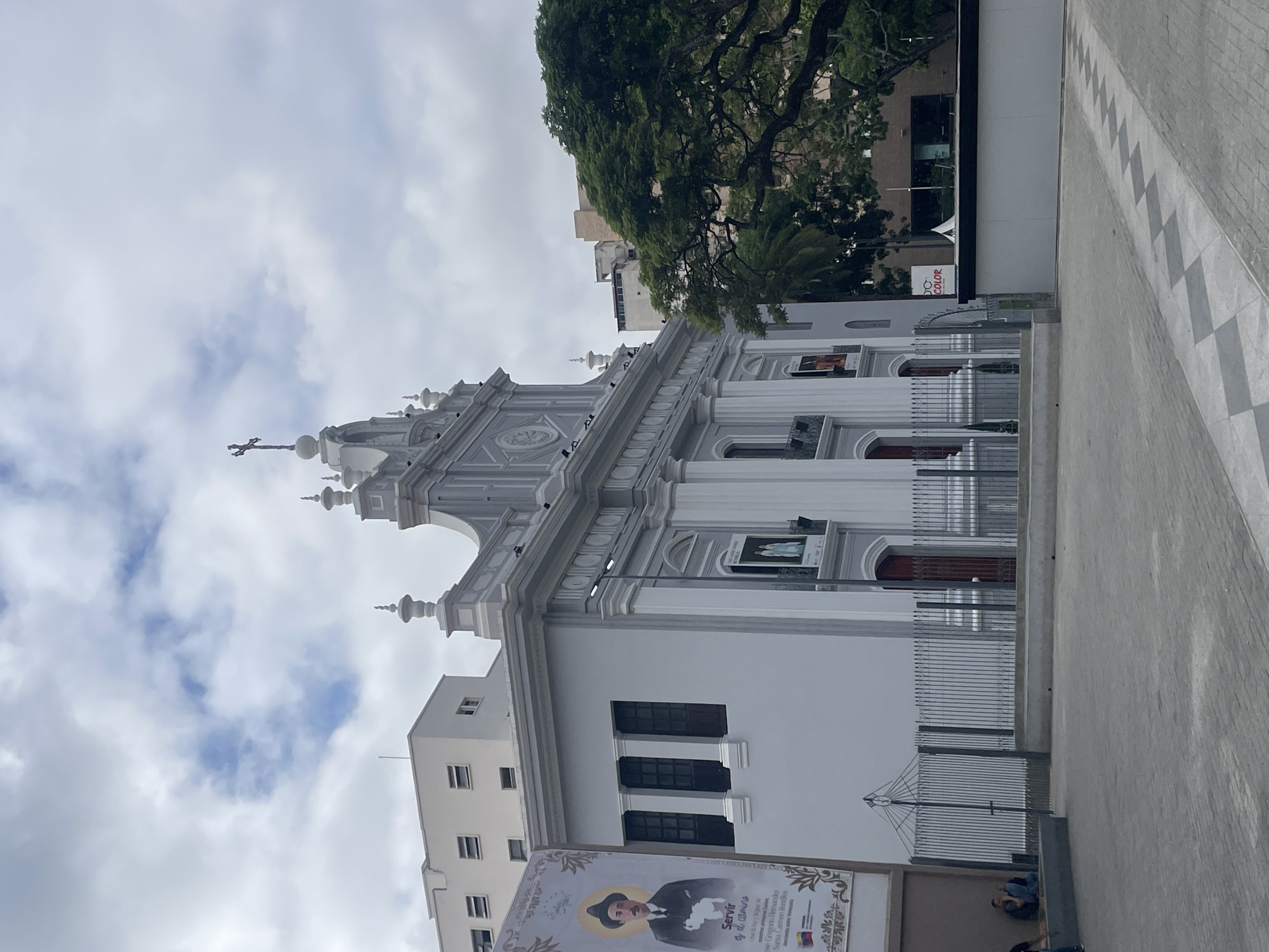 The image captures a portion of the Santa Capilla church in Caracas, Venezuela. The building is predominantly white and grey, showcasing a neoclassical architectural style. A prominent cross sits atop the structure. Adjacent to the church, a building with dark windows and a large poster featuring a portrait of a man in a hat is visible. The sky is overcast with cloudy skies. A large tree frames the upper portion of the image. A stone-paved walkway is in the foreground, suggesting an urban setting. People are sitting next to the poster.