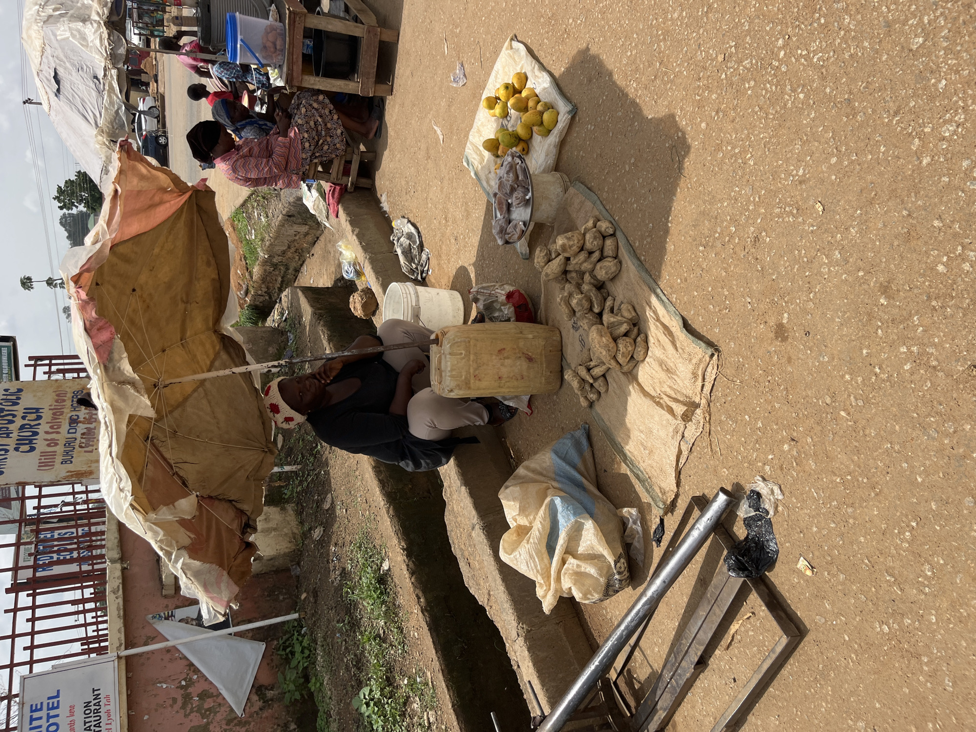 In Bukuru, Nigeria, a street vendor sits under a worn umbrella next to a drainage ditch. She is wearing a black top and light-colored pants, and appears to be talking on her phone. In front of her, on the ground, are several items for sale, including a large plastic container, a sack of produce, a pile of yams, and a tray of what appear to be packaged snacks. To the right, another vendor sits at a wooden stall, attending to customers, with a large blue bucket containing some goods. The background shows a street with cars and buildings, indicating a busy urban environment. The scene is typical of a market day in Nigeria, with vendors selling various goods on the roadside.