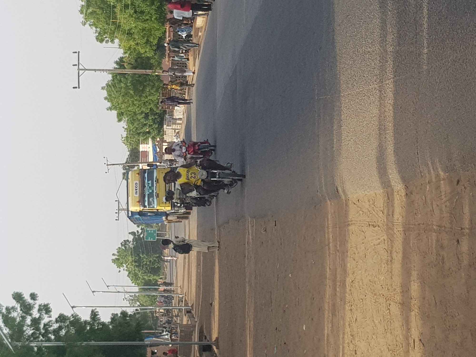 The image shows a street scene in Dabawa, Nigeria. The road is paved and runs horizontally through the frame. On the left side of the road, there are power lines and street lamps. Several people are gathered along the side of the road. Motorbikes and an IVECO truck are driving along the road. One man is walking on the side of the road. The sky is overcast, and there are trees lining the background.