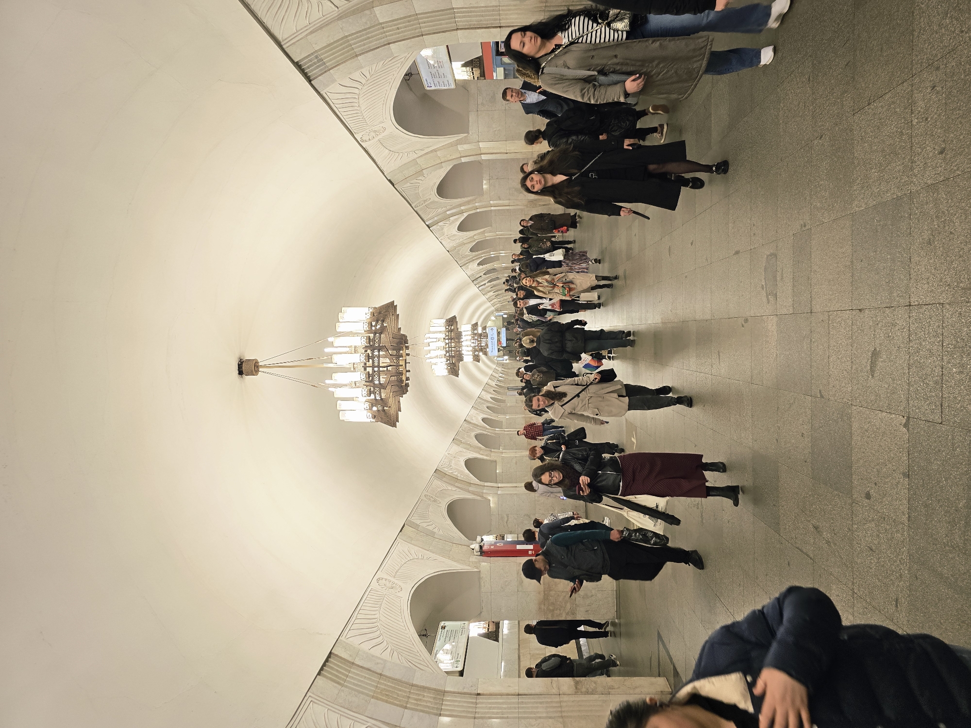 The image shows a long, vaulted hallway in a Moscow subway station. The hallway is lined with archways on either side. Ornate chandeliers hang from the ceiling. Many people are walking through the hallway. The floor is made of stone tiles. The walls and ceiling are primarily white, with ornate decorations above the archways. Some signs are visible within the archways. The scene is brightly lit.