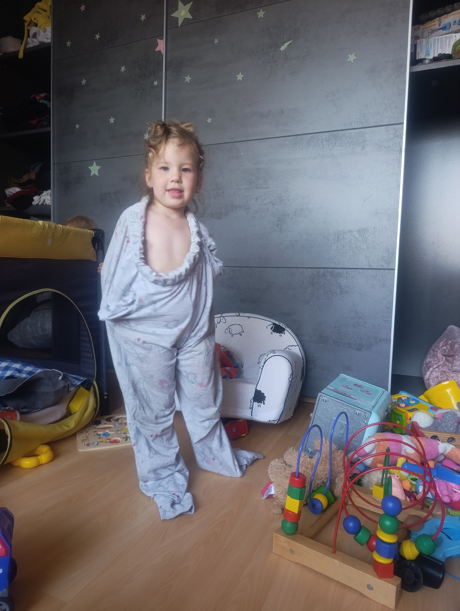 A young girl with blonde curly hair and a light-colored pajama set with a unicorn pattern is standing in what appears to be a bedroom or playroom. The room has a large gray wardrobe with star decals on the doors. To the left of the girl is a yellow and black playpen with a blue and white checkered blanket inside. In front of the wardrobe, there is a small white armchair with black sheep designs. The floor is made of light wood. Various toys are scattered around, including a wooden bead maze, a stuffed bear, a toy car, and a blue toy chest. The girl is wearing oversized pajamas, with the top part pulled up around her neck and shoulders, and the pants pooling around her feet. The overall impression is that of a child playing dress-up. The image does not provide any specific information about the location in Olomouc, Czechia.