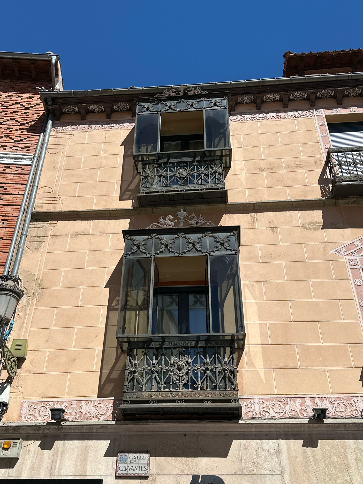 The image captures an upward view of the facade of a building on Calle de Cervantes in Segovia, Spain. The building features a light peach-colored stucco exterior with a pattern of lighter peach-colored rectangular blocks. Two ornate metal balconies with intricate wrought-iron railings are visible on the upper floors. The balconies have dark metal frames and glass panels. Above and below the balconies, there are decorative friezes. A red brick building with a visible drainpipe is to the left, and a portion of another building with a balcony and grey roller shutters is to the right. The sky is clear and blue. A street sign reading "CALLE DE CERVANTES" is mounted on a lower section of the building.