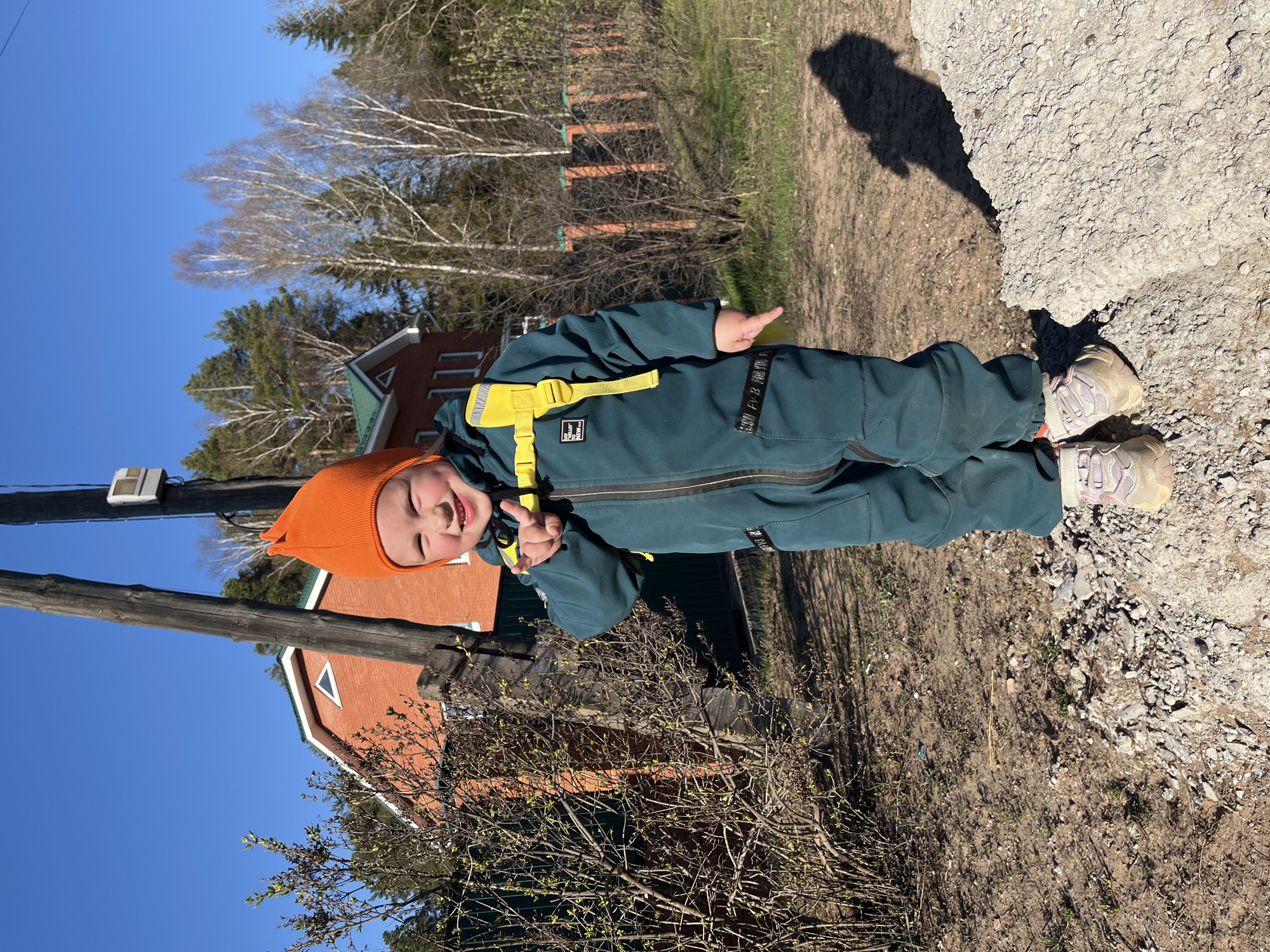 A young child, dressed in a dark green jumpsuit and orange hat, stands outside on a sunny day. The child is holding up two fingers in a "peace" sign. The location appears to be a semi-rural area with a red brick building and bare trees in the background. A utility pole is visible to the left. The ground is dirt and gravel. Based on the information provided, it is plausible this location is in or near the city of Chaikovsky, Russia.