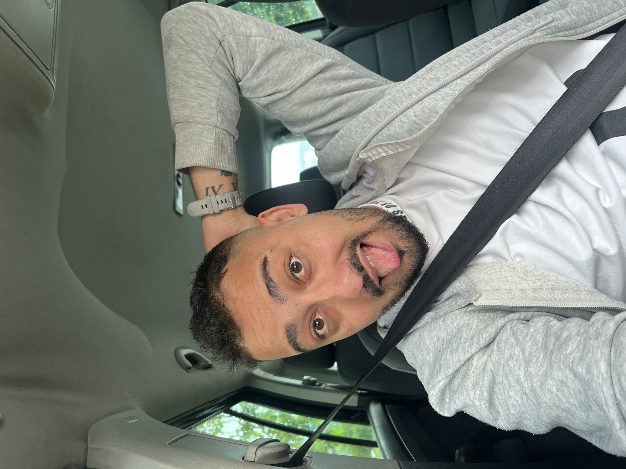 A selfie of a man in a car, angled such that the man's head is at the bottom of the frame. The man has brown eyes, short dark hair, and a trimmed beard. His tongue is sticking out. He is wearing a white shirt, a light gray zip-up hoodie, and a gray smartwatch with a dark-colored tattoo on the wrist. A black seatbelt crosses diagonally across his body. The car interior is visible, including the roof, dashboard, and window showing greenery outside. The location cannot be determined to be in Касапите, Bulgaria based on visual data.