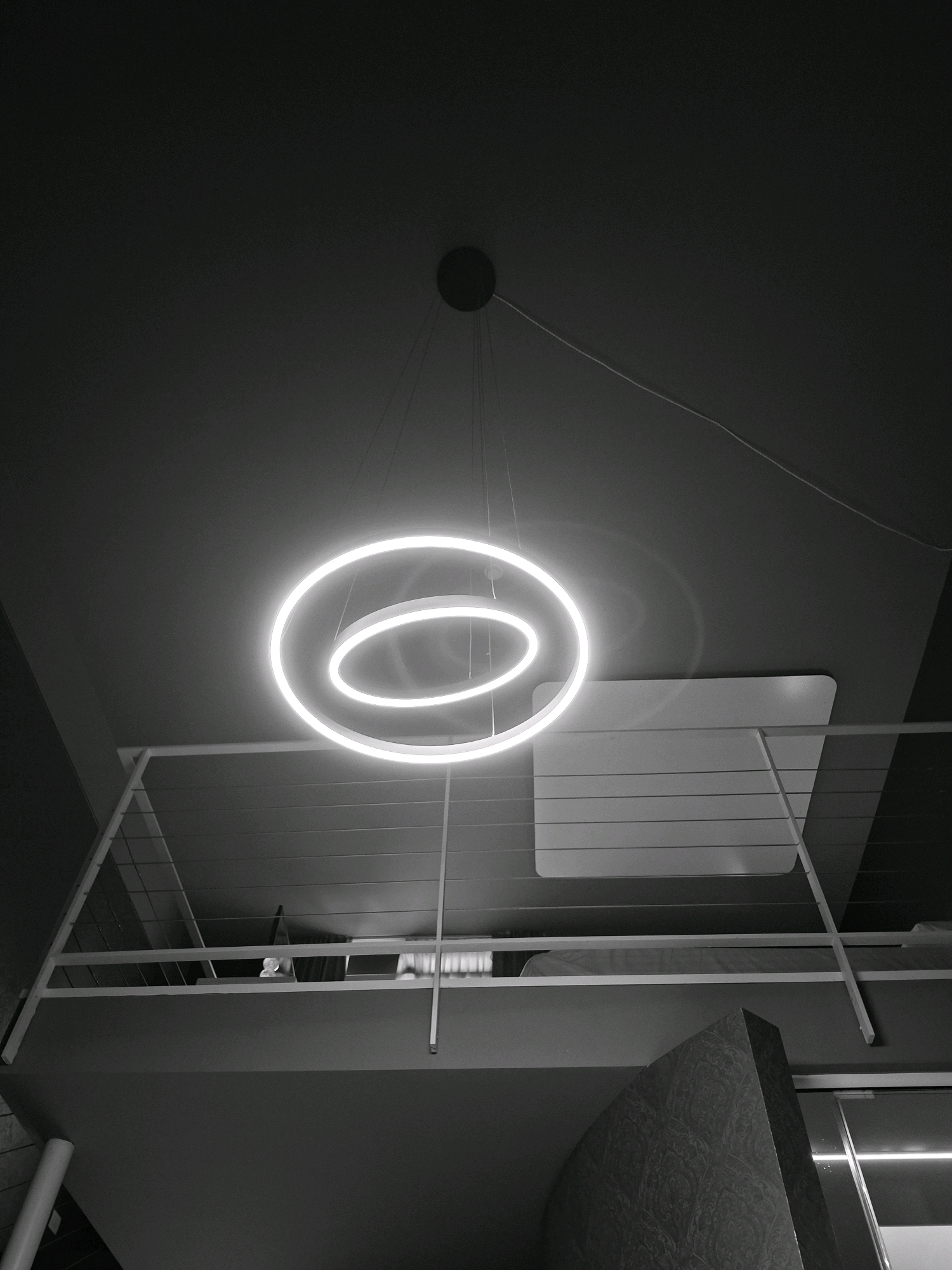 The image shows a modern, dimly lit interior space, likely a residential or commercial building. A prominent feature is a circular, illuminated pendant light fixture hanging from the ceiling. Below the light, a contemporary mezzanine level is visible, characterized by a white metal railing and horizontal slats on its underside. The overall aesthetic is minimalist and sleek, with a grayscale color palette. The scene is devoid of any people. There is no specific information within the image to identify its location within the city of Olgiate Olona, Italy.