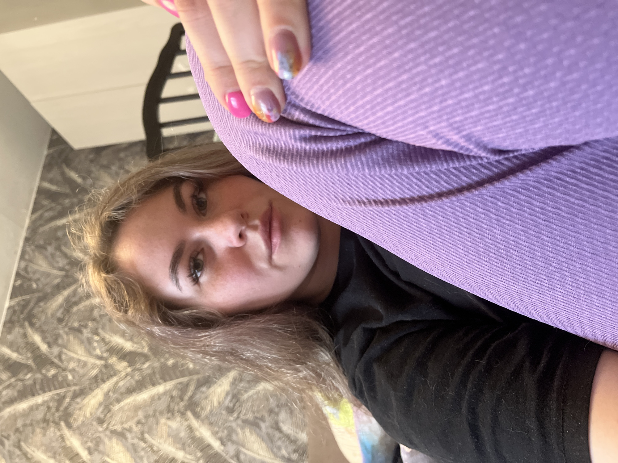 A close-up, selfie-style portrait captures a young woman resting indoors, looking directly at the camera. Her face, positioned centrally in the lower half of the frame, reveals her left eye, nose, and lips. She has light brown/blonde hair framing her face, and visible freckles dot her nose and cheeks. Her expression is neutral and calm.
A hand with distinctively painted fingernails is prominent in the upper right quadrant. One nail is a solid bright pink, while others feature an iridescent, multi-colored design with hues of pink, orange, blue, and silver. This hand appears to be holding or resting on a large piece of lavender or light purple fabric, which occupies a significant portion of the image. The fabric has a distinct ribbed or corduroy-like texture. The woman is wearing a black garment, likely a t-shirt or top, visible beneath the purple fabric.
The background shows a subtly patterned surface, possibly wallpaper or a textile, with abstract, swirly designs in muted tones of gray and brown. A dark, decorative piece of furniture, resembling the back of a chair, is partially visible in the upper left background. The overall impression is one of relaxation in a personal, domestic setting. No text is visible in the image.