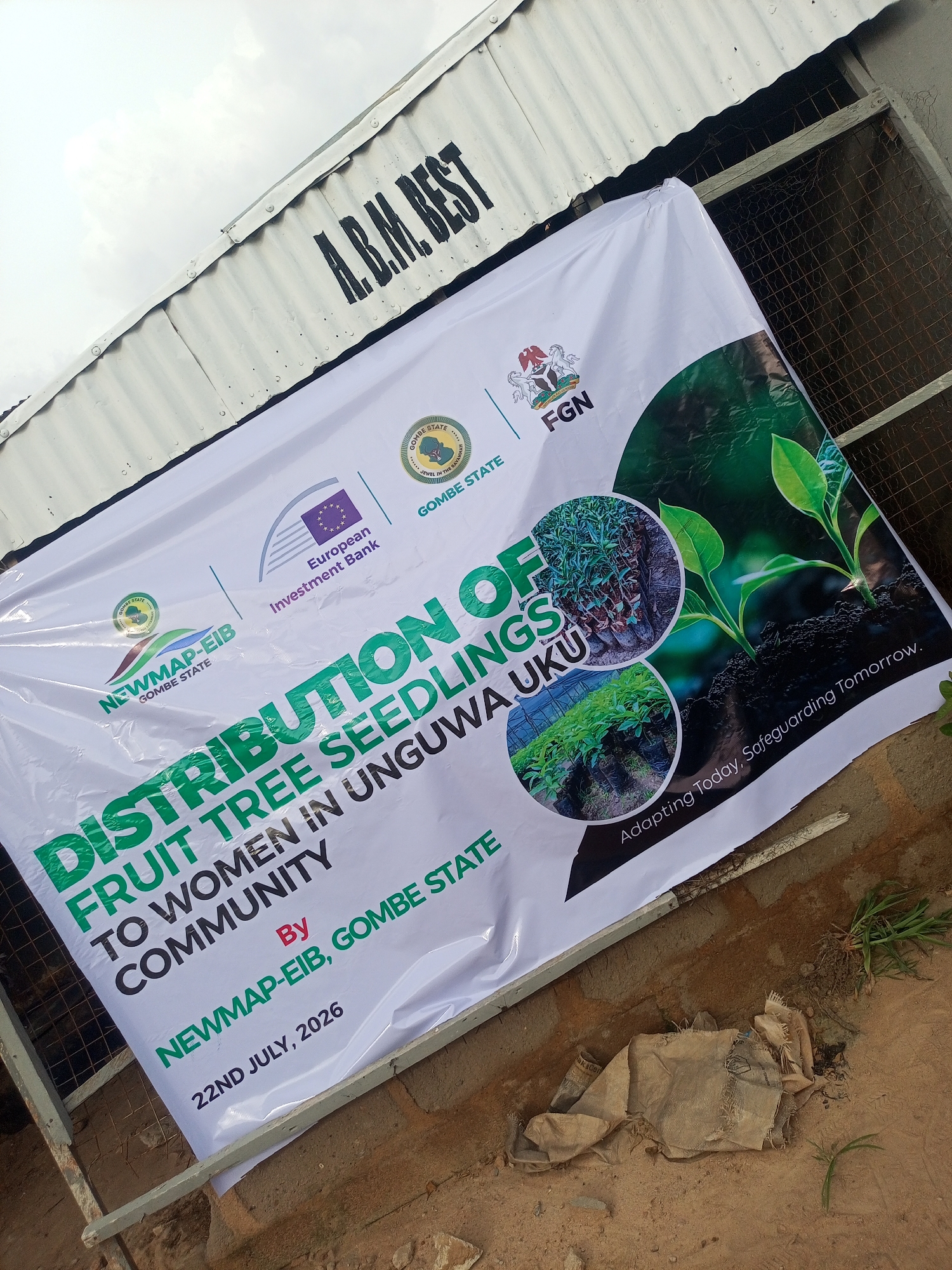 An outdoor, slightly tilted medium shot captures a white banner advertising the "DISTRIBUTION OF FRUIT TREE SEEDLINGS TO WOMEN IN UNGUWA UKU COMMUNITY BY NEWMAP-EIB, GOMBE STATE". The banner is partially unfurled and draped over a metal fence, leaning against a corrugated metal structure with "A.B.M. BEST" stenciled in black on its upper portion. The banner features several logos, including the Gombe State emblem, the European Investment Bank logo, and the Nigerian coat of arms with "FGN" beneath it. Two circular images are printed on the banner: one showing rows of young plants in bags, and another depicting two seedlings growing in dark soil. Text on the banner also reads "Adapting Today, Safeguarding Tomorrow." and "22ND JULY, 2026". The environment appears to be a dusty, outdoor area with sparse vegetation and some scattered debris, including a dirty burlap sack. The sky is overcast.