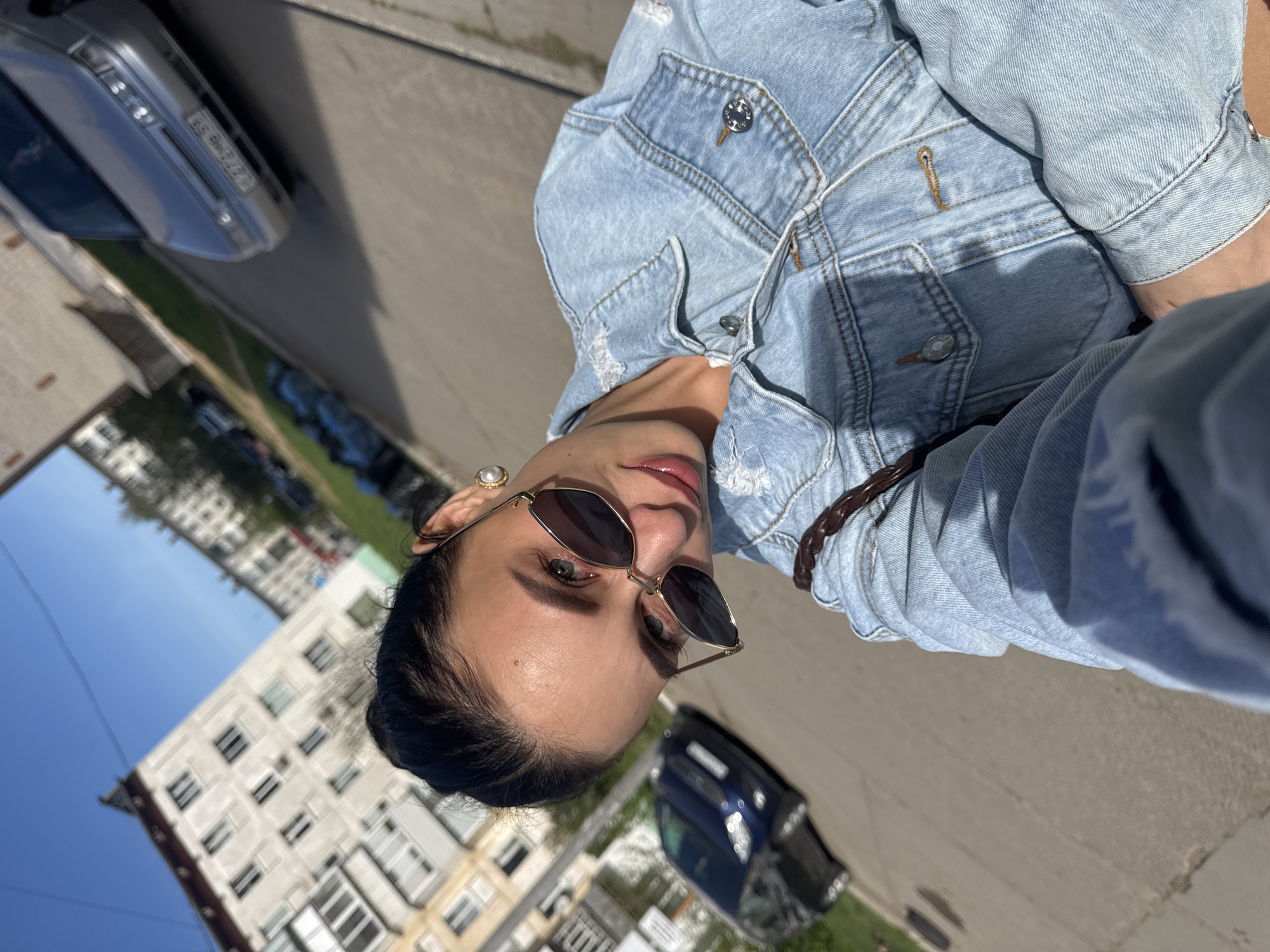 A selfie captured in Chaikovsky, Russia, reveals a woman donning a light blue denim jacket and sunglasses. She is positioned on a street in the city, where a multi-story apartment building is visible in the background. Two cars, a silver sedan and a dark-colored vehicle, are parked along the side of the road. The woman's hair is dark, and she is wearing a pearl earring. The scene is illuminated by sunlight, suggesting a bright day.
