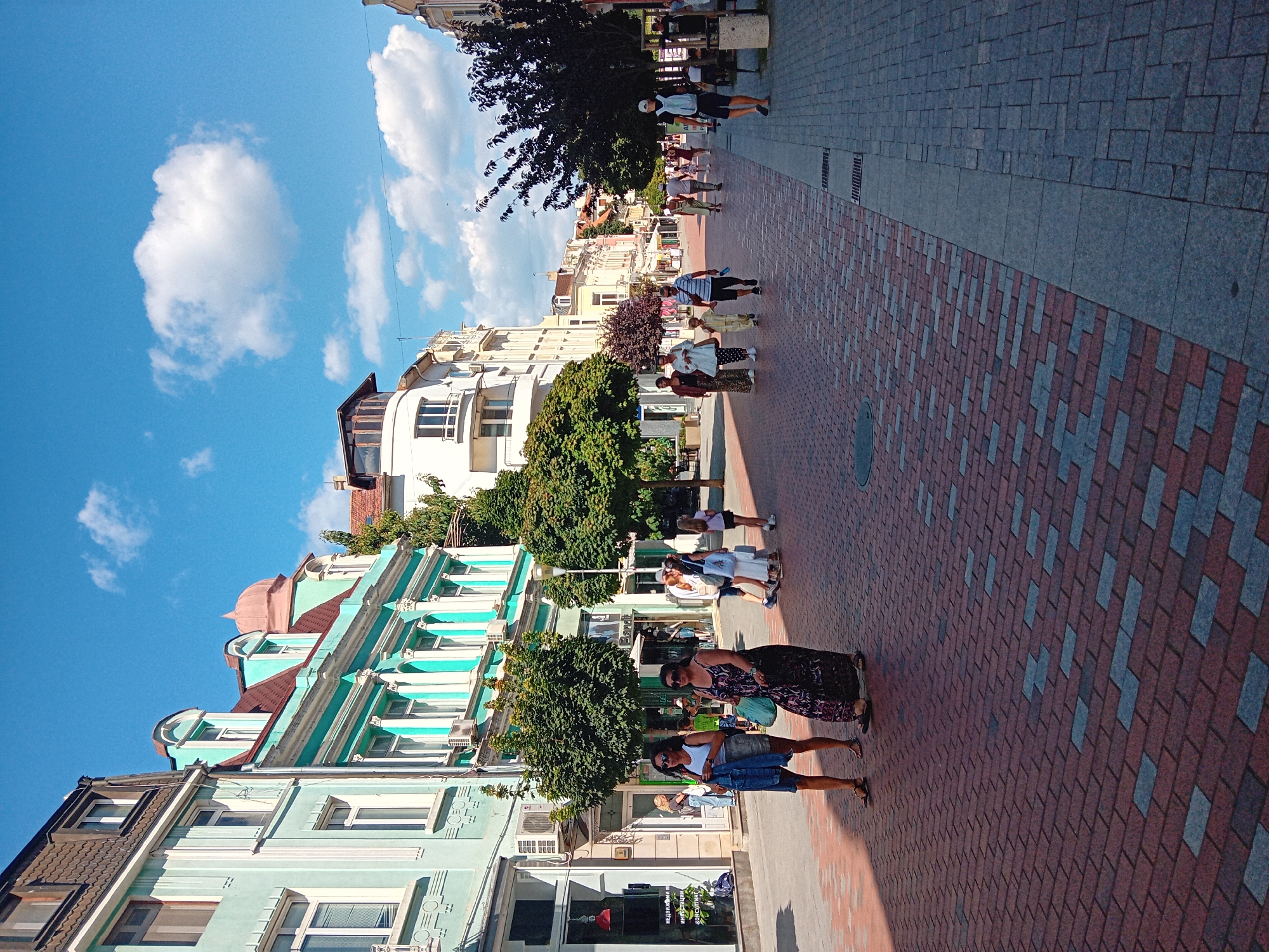 The image depicts a pedestrian street in Varna, Bulgaria, likely on a sunny day with scattered clouds. The street is paved with red and gray bricks, forming a textured walkway. On the left side of the street, there are several buildings with ornate facades, painted in light blue and white with turquoise accents. These buildings feature multiple stories, arched windows, and decorative cornices. Shops with signs are visible at ground level.
On the right side of the street, there are more buildings in the background, painted in lighter colors, including white and yellow. Trees are planted along the sides of the street, providing shade.
Several people are walking along the street. In the foreground, two women are walking towards the viewer, one in a white tank top and denim shorts, carrying a denim jacket, and the other in a long, patterned dress. Further down the street, a group of people, including adults and children, are seen walking in various directions. A man wearing a white cap and backpack is walking away from the viewer on the right side of the street. The overall atmosphere appears to be that of a bustling, lively urban environment.
