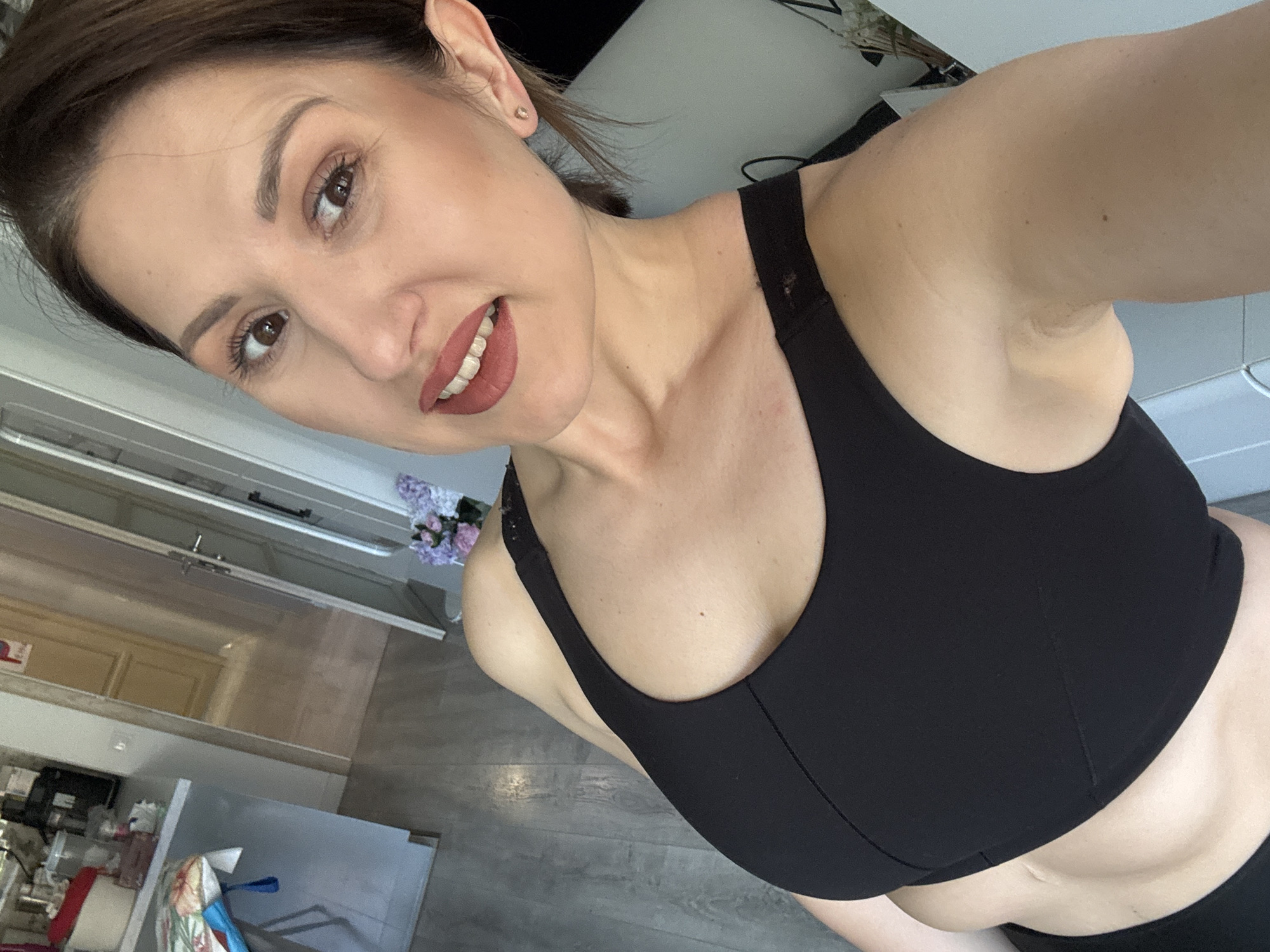 A medium shot, taken at an angle, shows a light-skinned woman with short, dark brown hair and brown eyes, smiling at the camera. She is wearing a black sports bra. Her lipstick is a shade of reddish-brown. A small stud earring is visible in her left ear.
Behind her, a kitchen area is visible. To the left, there are white cabinets and drawers with silver handles. On the counter, there is a metal sink and miscellaneous items, including a red can, a white paper towel roll, and a colorful cloth. Above the cabinets, there is a translucent window or door.
In the background, there are light-colored wooden floors. To the top right, there are blurry white objects.
The location in the city of Sofia, Bulgaria cannot be determined from the image.